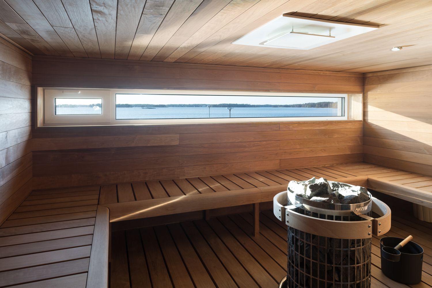 A wooden sauna interior with tiered benches, a stone heater, and a horizontal window looking out over water.