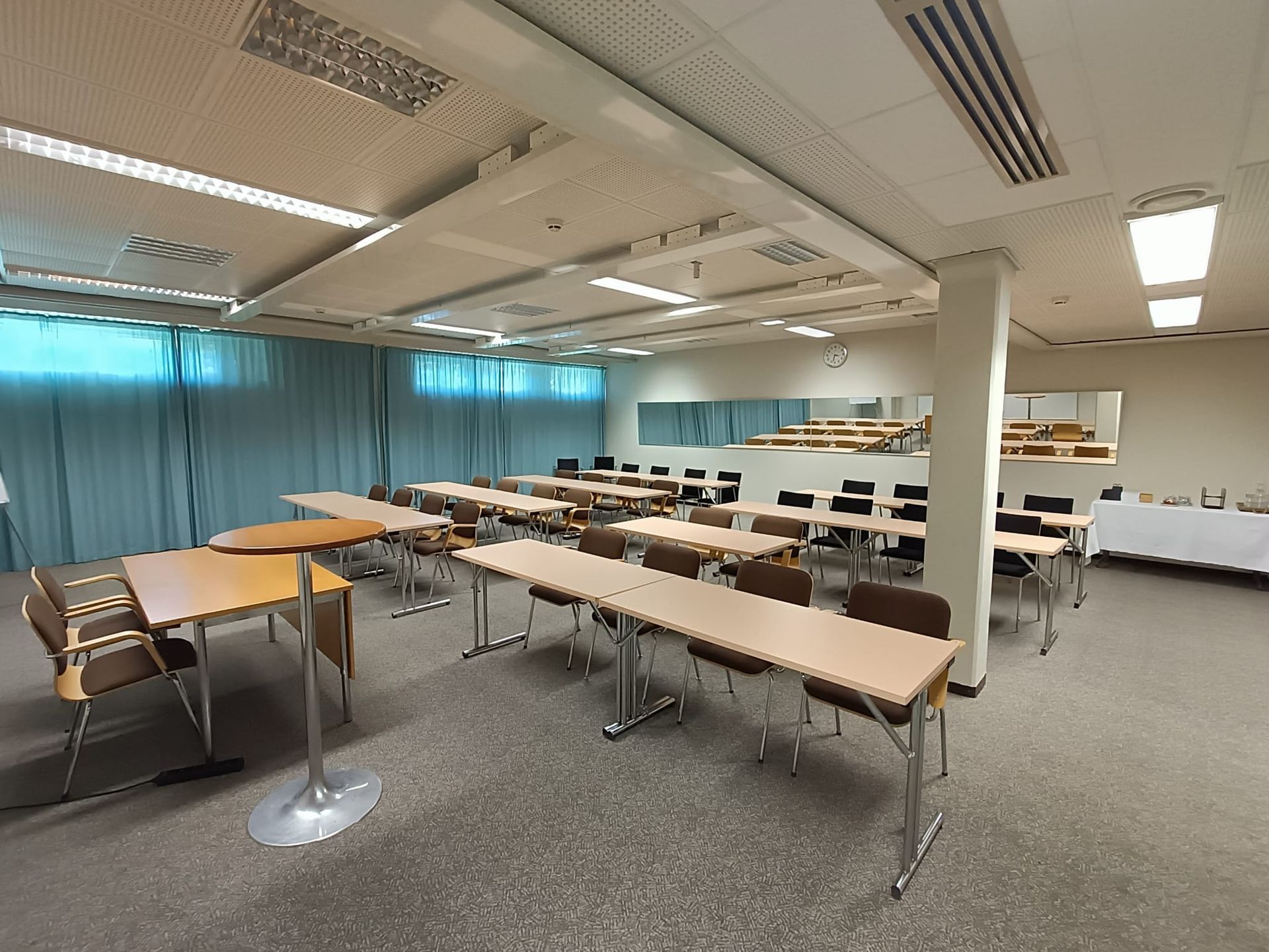 A large, empty conference room with rows of tables and chairs, blue curtains, and fluorescent lighting.
