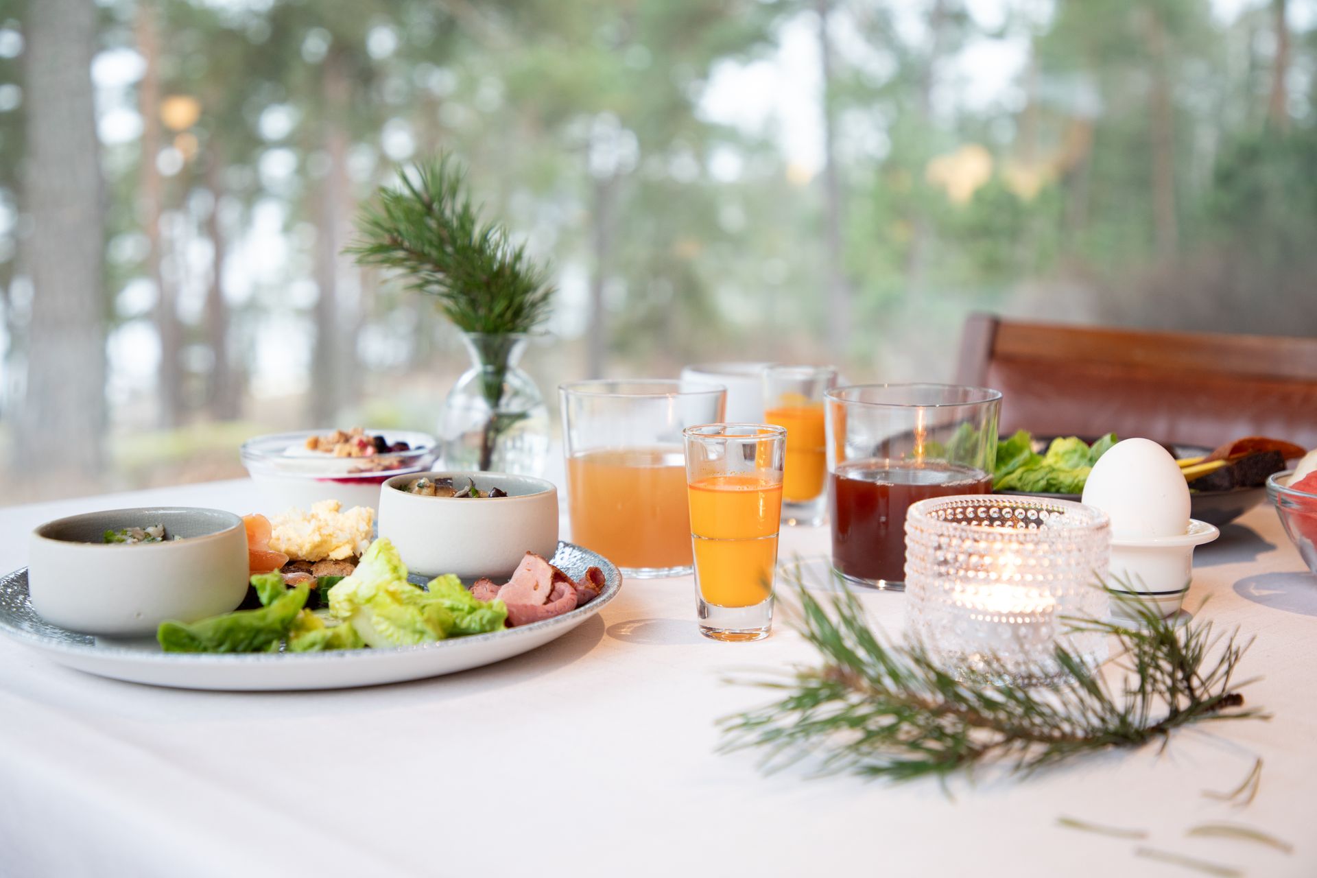 A breakfast spread with small bowls of food, juices, a soft-boiled egg, and a lit candle on a white table near a window.