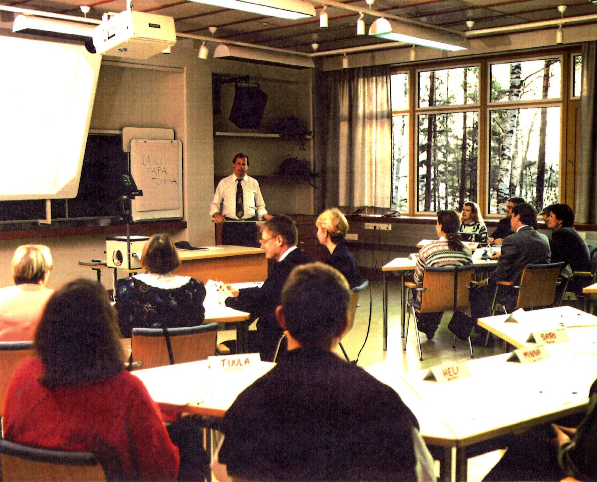 A speaker stands at the front of a brightly lit classroom as a group of people sit at tables facing them.