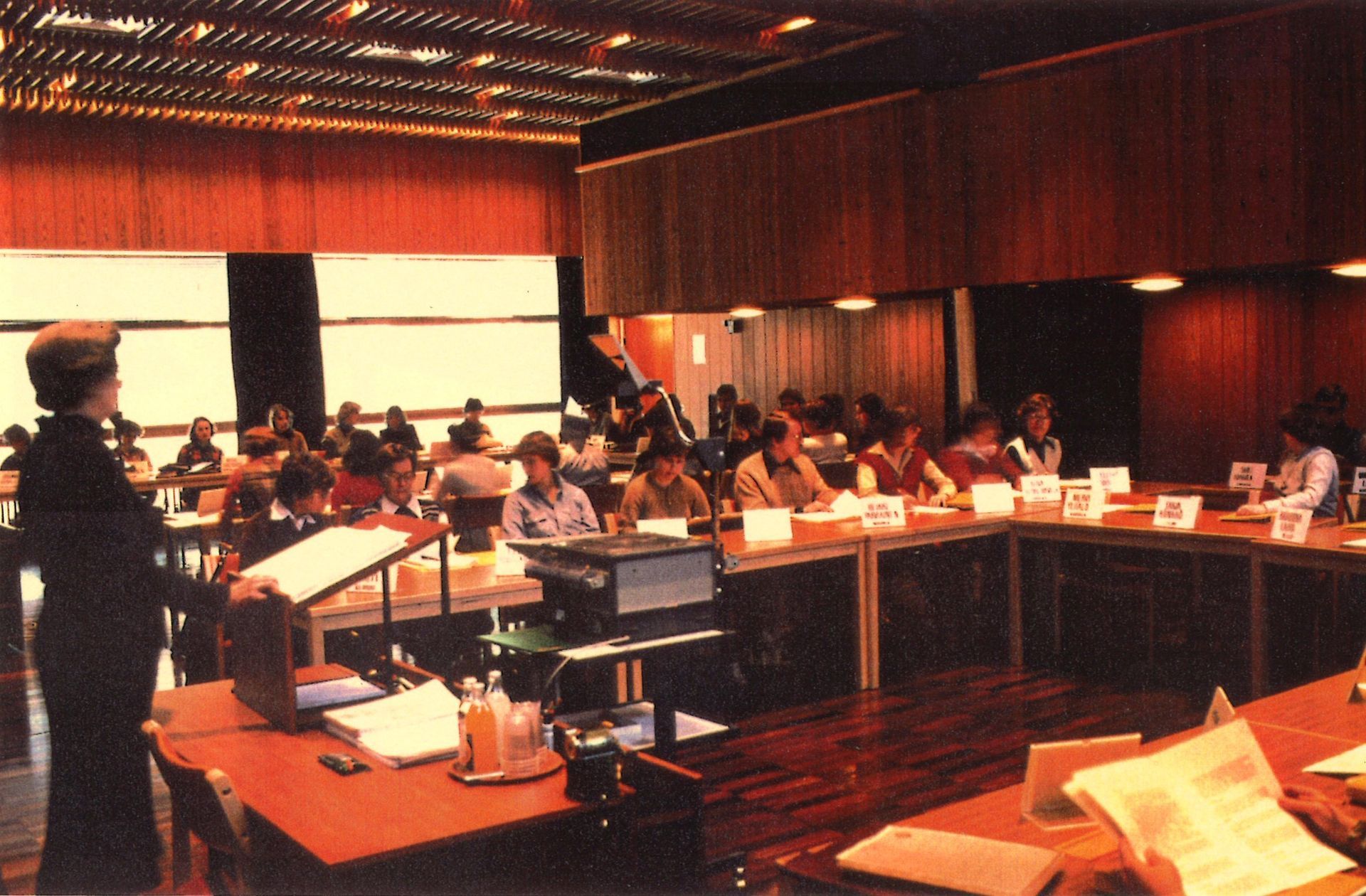 A person stands at a podium addressing a group of people seated around a U-shaped table in a wood-paneled conference room.