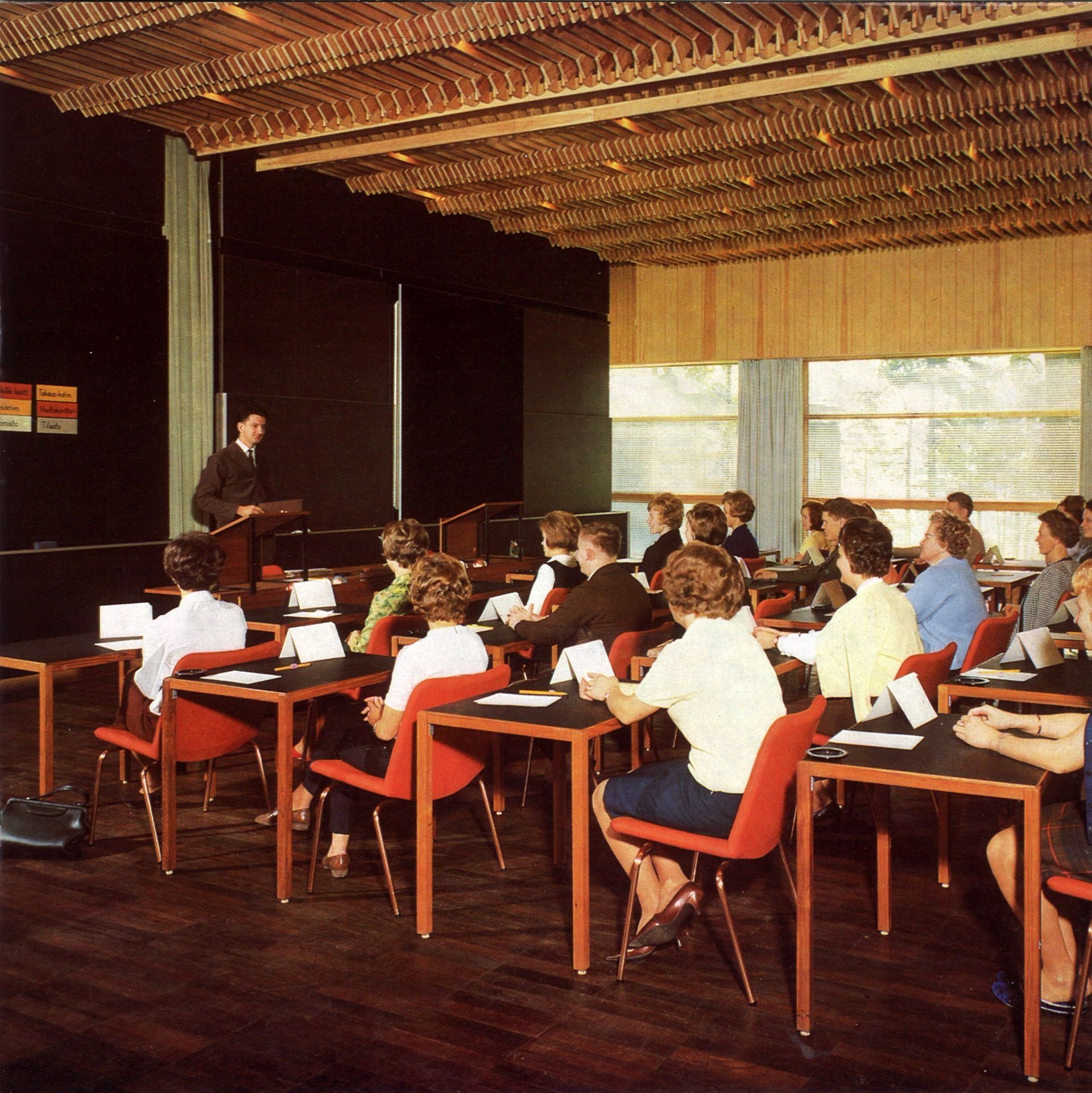 A speaker stands at a podium addressing an audience seated at desks in a modern, wood-paneled classroom with red chairs.