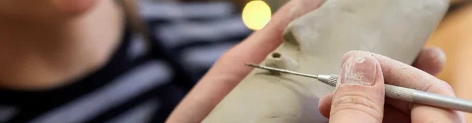 A person uses a metal sculpting tool to carve detail into a light-colored clay figure.