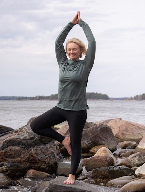 A person balancing in a yoga tree pose with hands pressed together overhead, standing on rocks by a body of water.