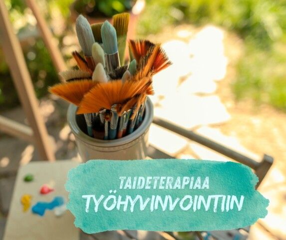 Paintbrushes in a container next to a paint palette with a teal watercolor banner reading "Taideterapiaa työhyvinvointiin".