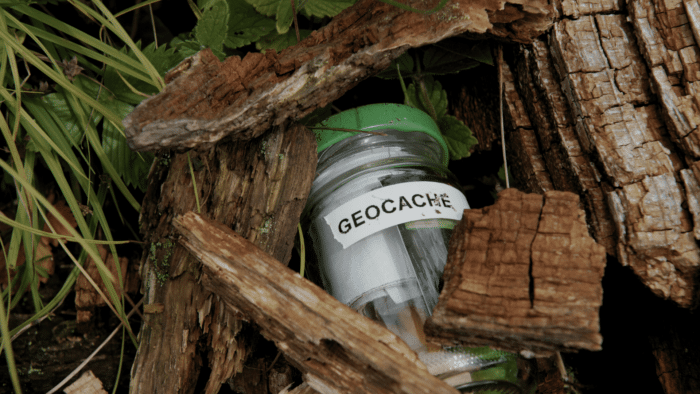 A small clear jar labeled "GEOCACHE" with a green lid, hidden among pieces of rough tree bark and greenery.