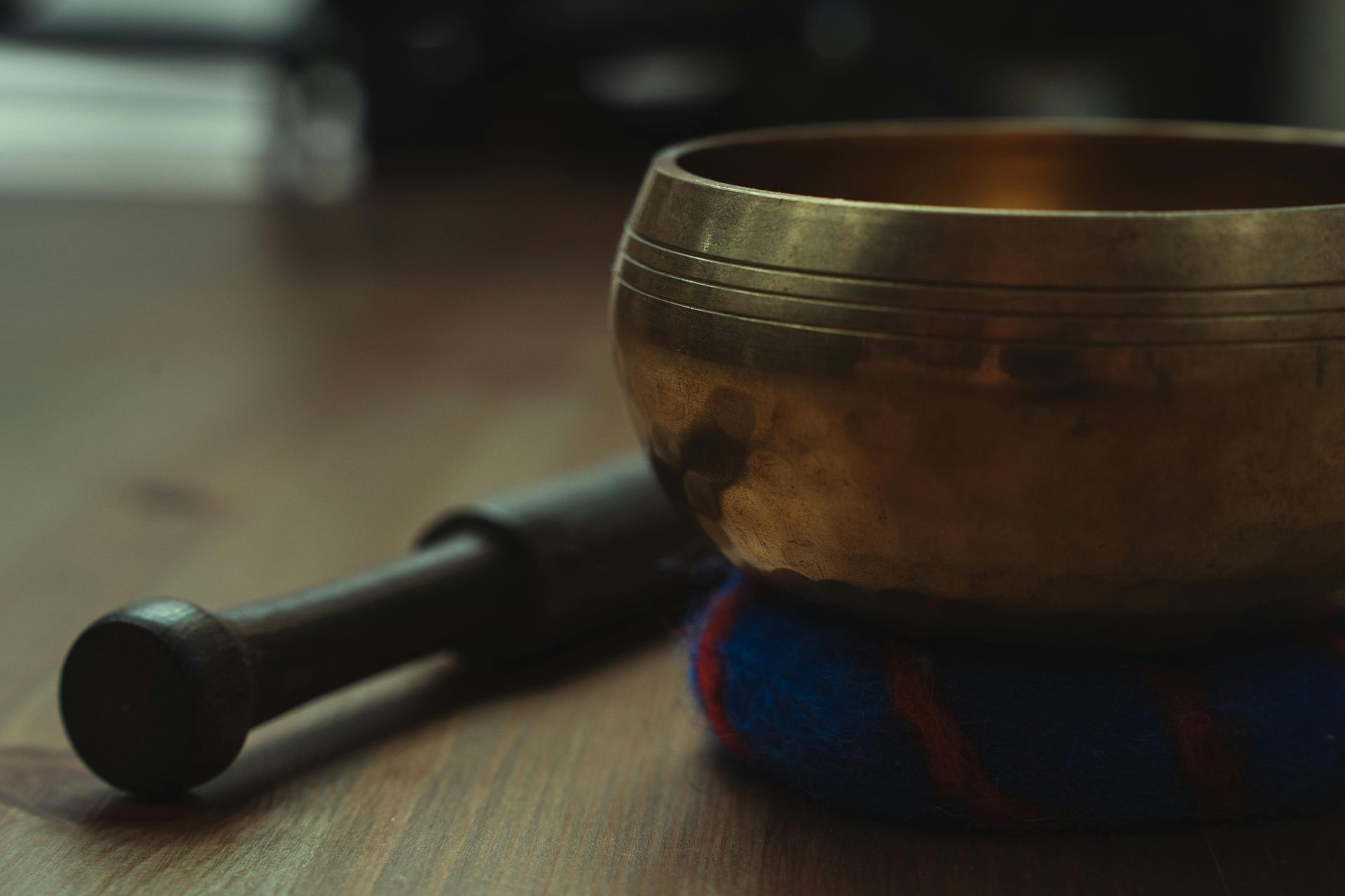 A metallic singing bowl resting on a blue and red cushion, with a wooden mallet lying on a wooden surface beside it.