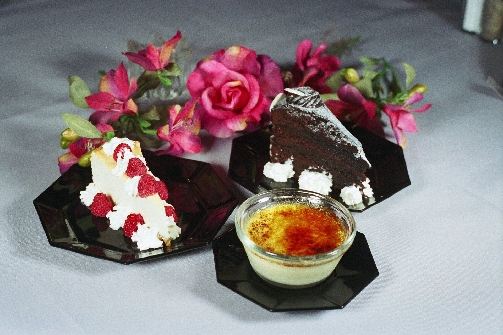 Desserts on black plates with flower arrangement: cheesecake, chocolate cake, and crème brûlée.