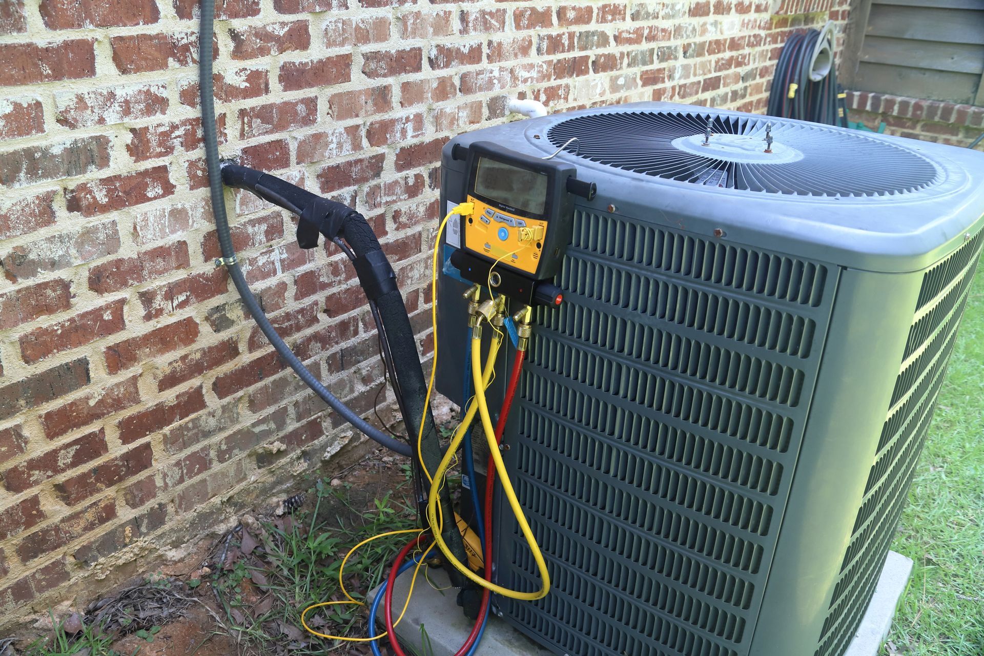 HVAC technician testing residential outdoor air conditioner with gauges beside brick house exteriors.