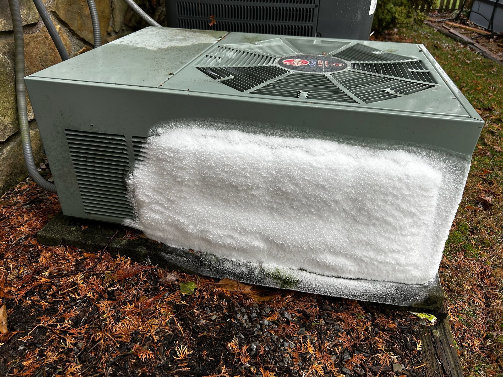 Outdoor AC unit with ice buildup on the side. The unit is light green, sitting on a dark base with dry leaves around.