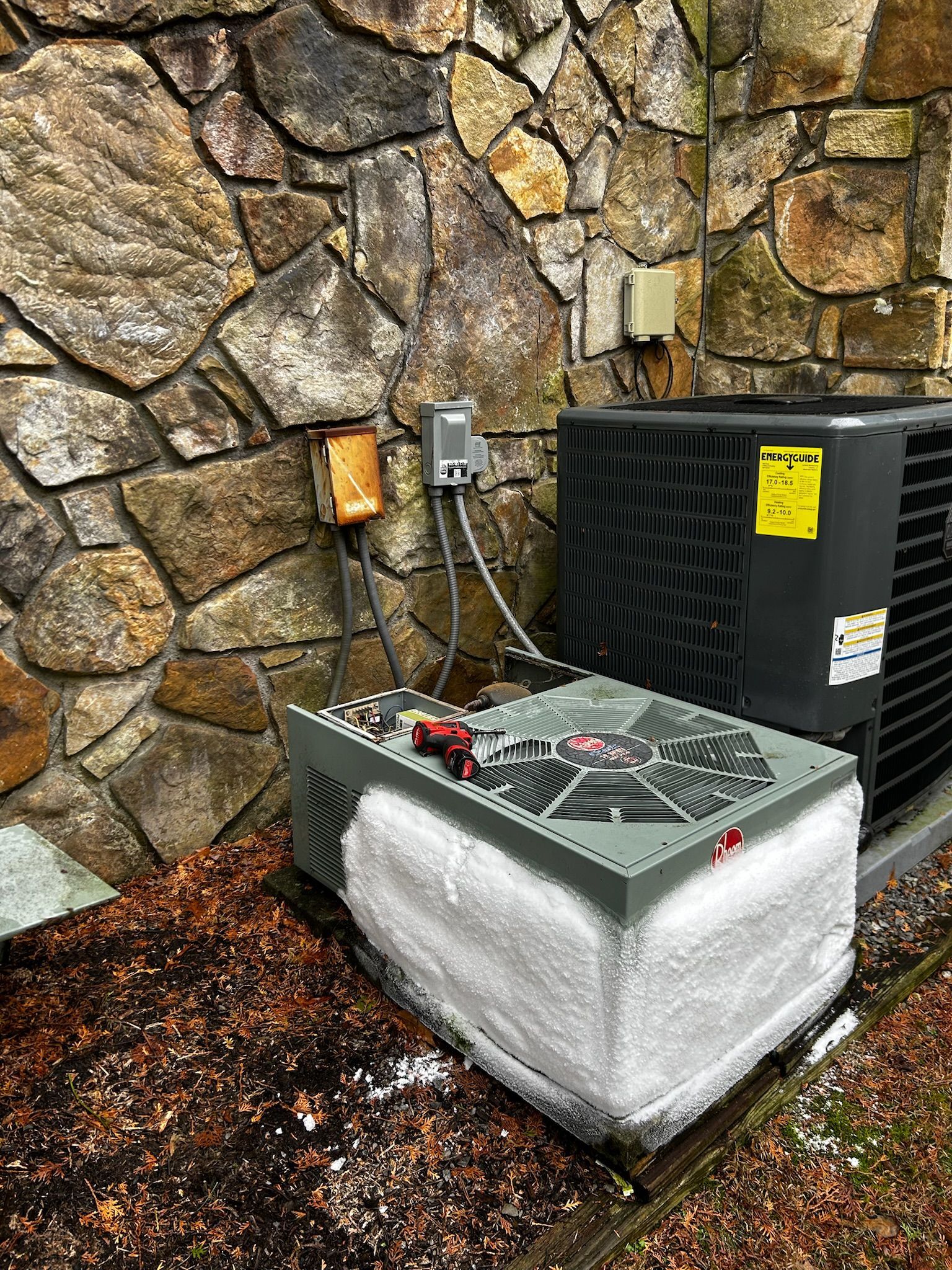 Outdoor air conditioning unit beside a stone wall, with electrical boxes and wet ground nearby.