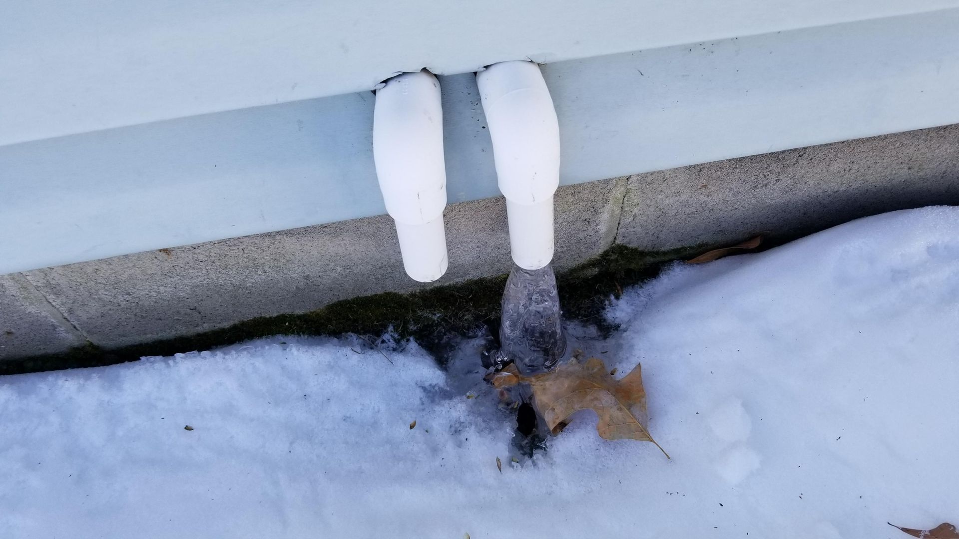 White downspouts with melting ice, releasing water onto snow-covered ground.