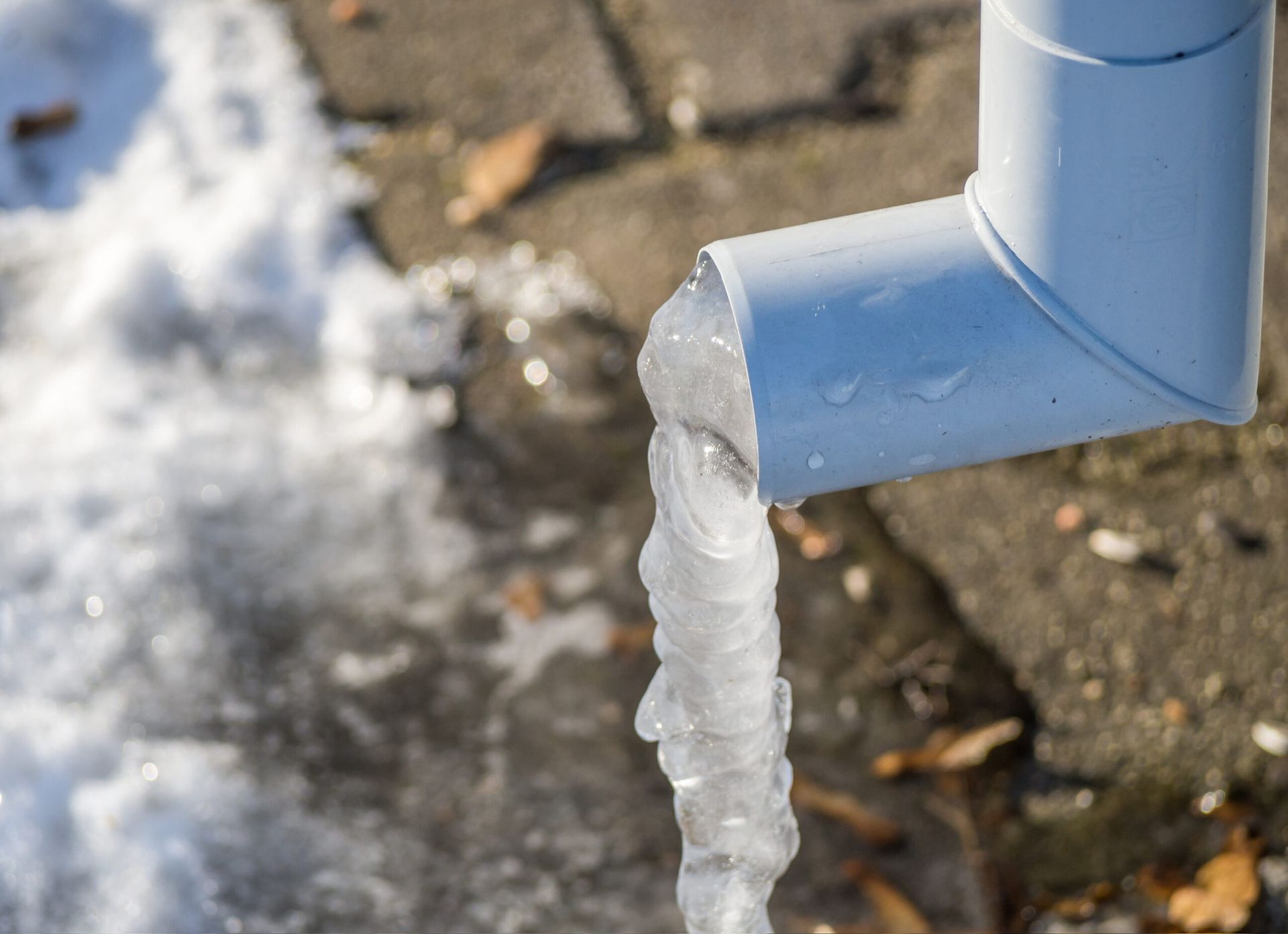 White downspout with ice hanging from the end, snow and pavement visible.