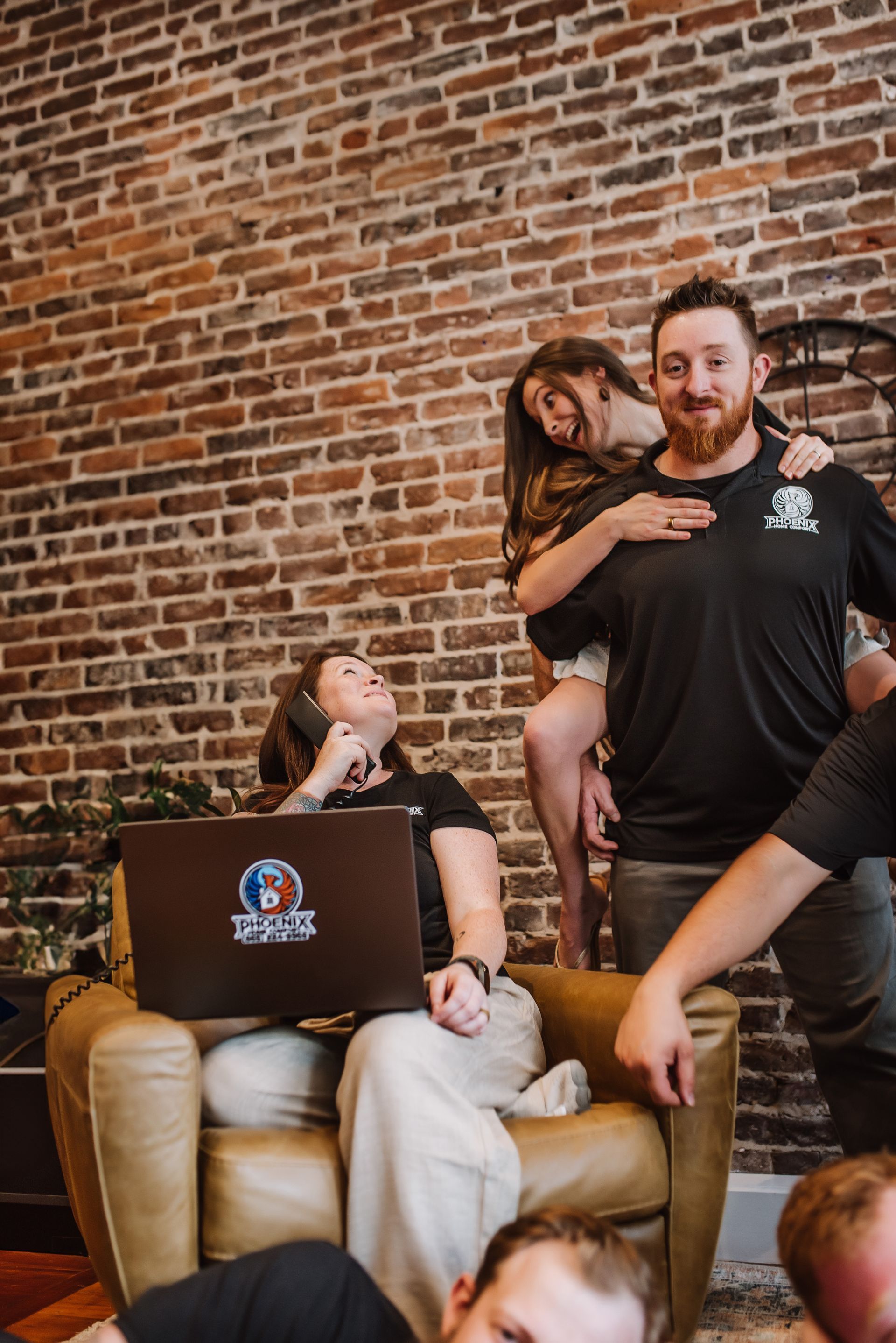 People relaxing in chairs against a brick wall, with one person on another’s shoulders and a laptop open.
