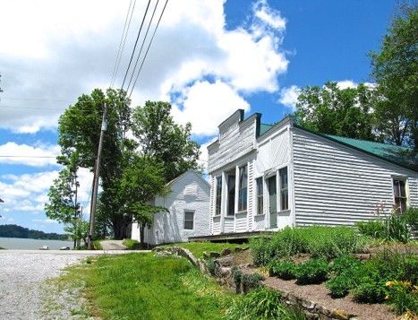 White waterfront house with green roof beside a gravel road, trees, and blue sky with clouds