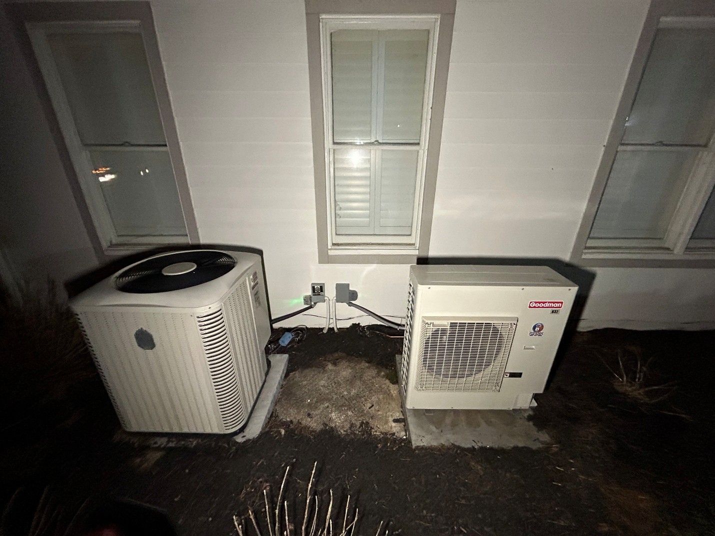 Two outdoor HVAC units beside a house wall beneath two windows at night. Two outdoor HVAC units beside a house wall beneath two windows at night.