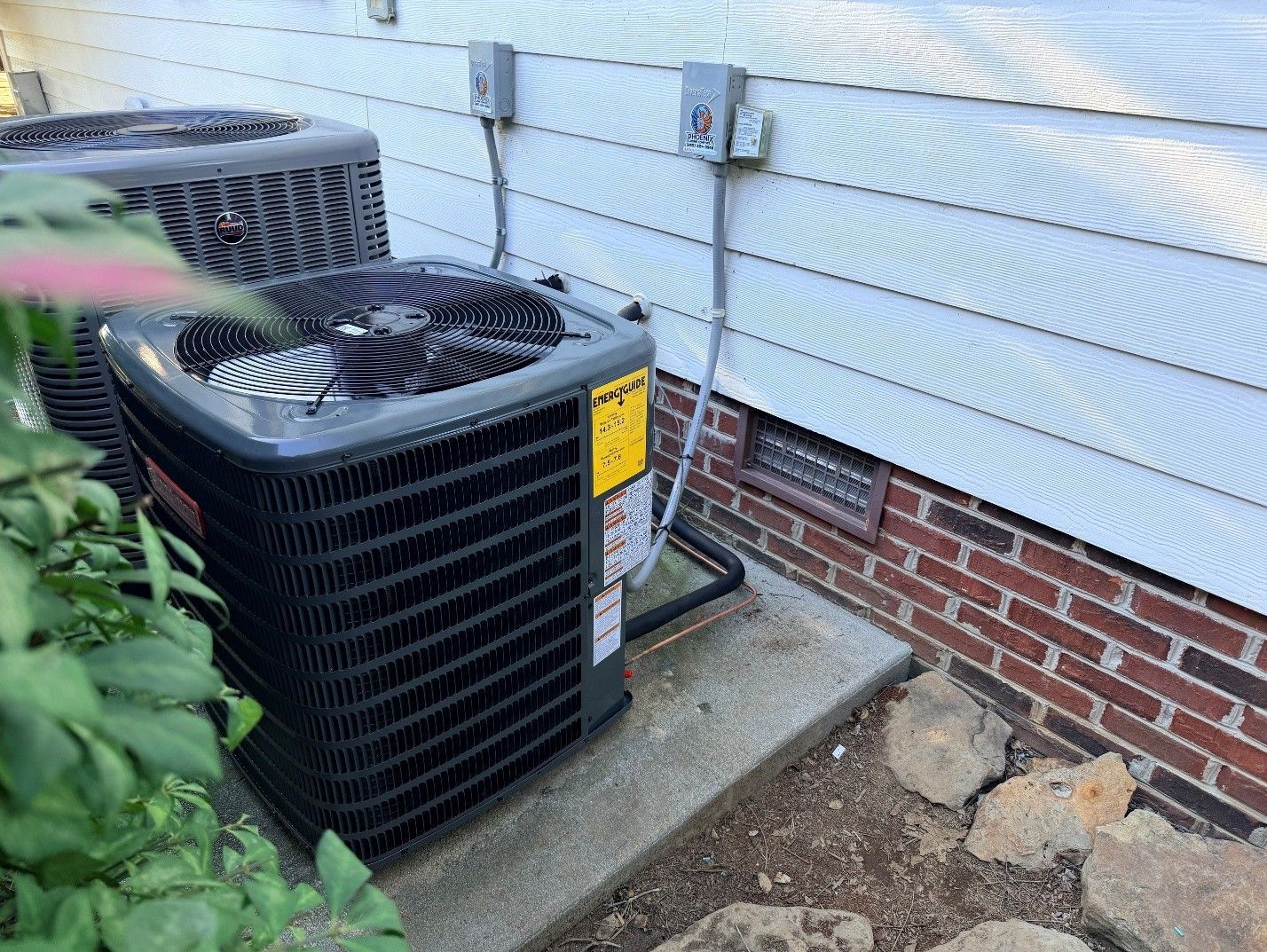Outdoor air conditioner units beside a house wall on a concrete pad with gravel and plants nearby