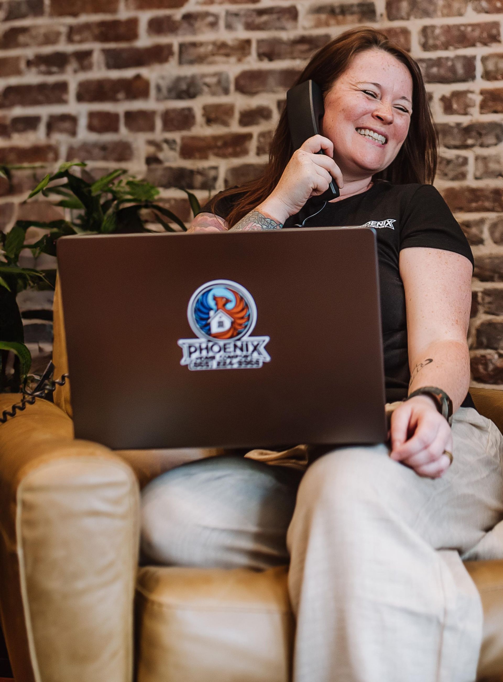 Smiling woman on a tan couch using a laptop in front of a brick wall