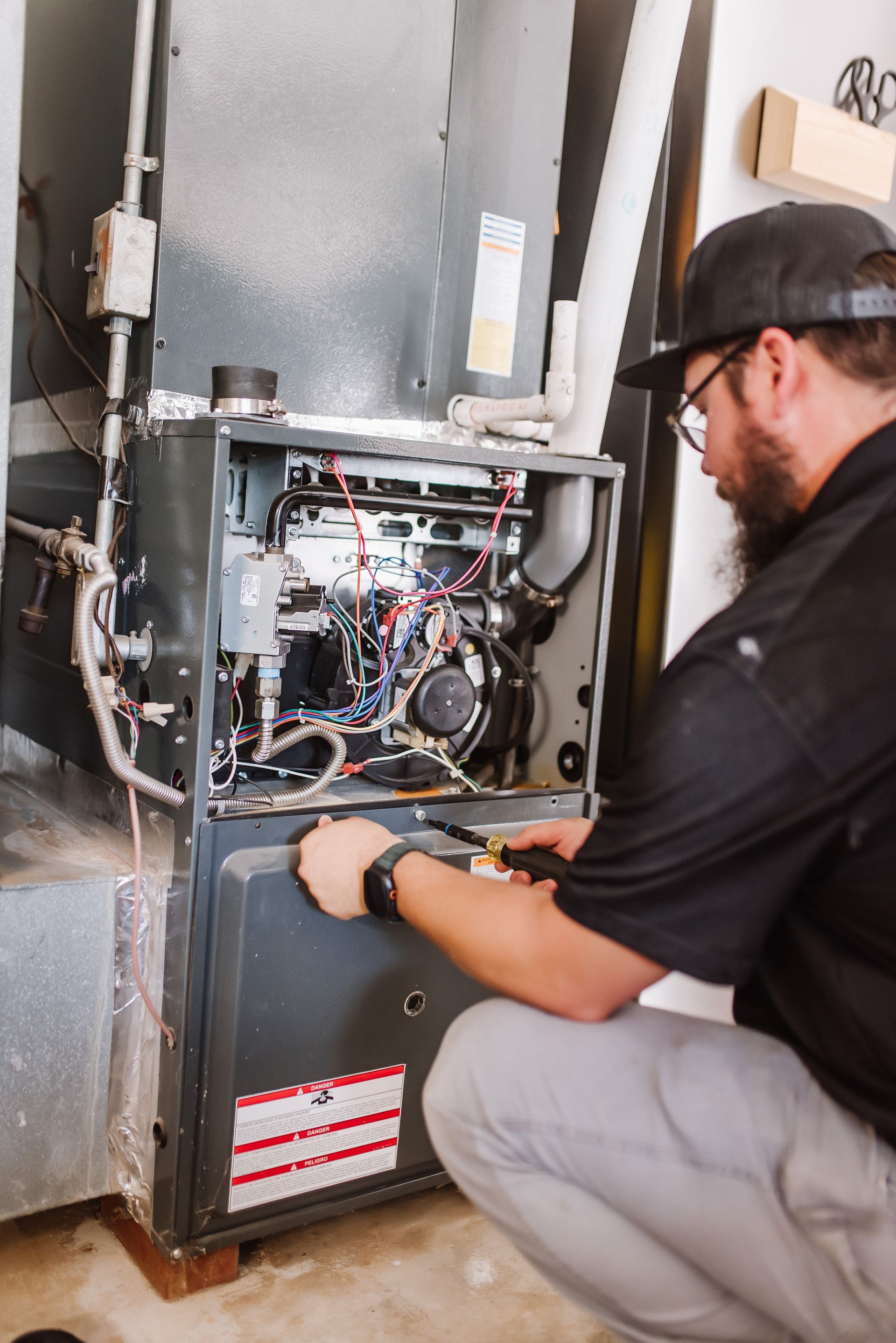 Technician kneeling beside an open furnace panel, inspecting exposed wiring and components.