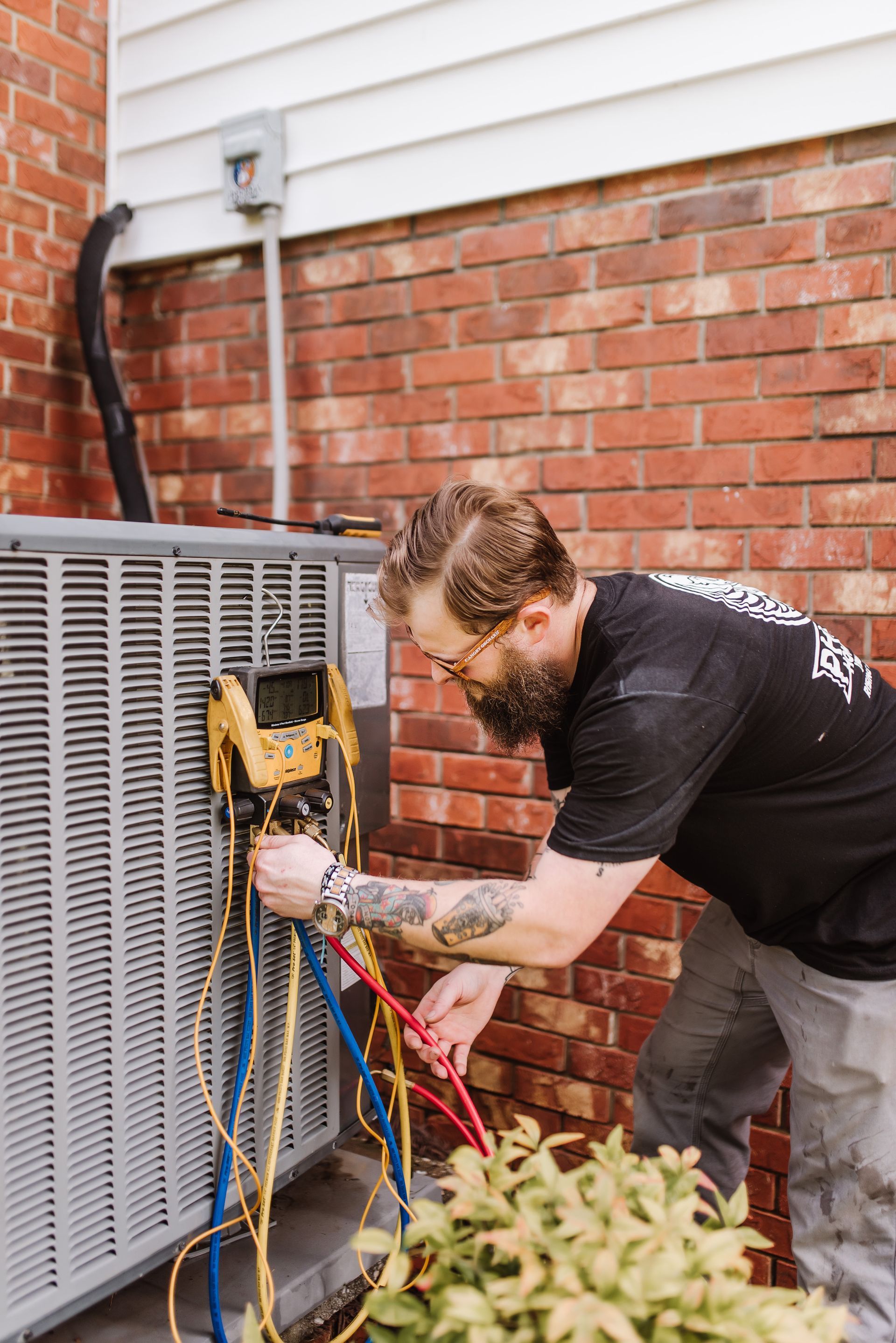 Technician servicing an outdoor HVAC unit beside a brick wall, using gauges and tools.