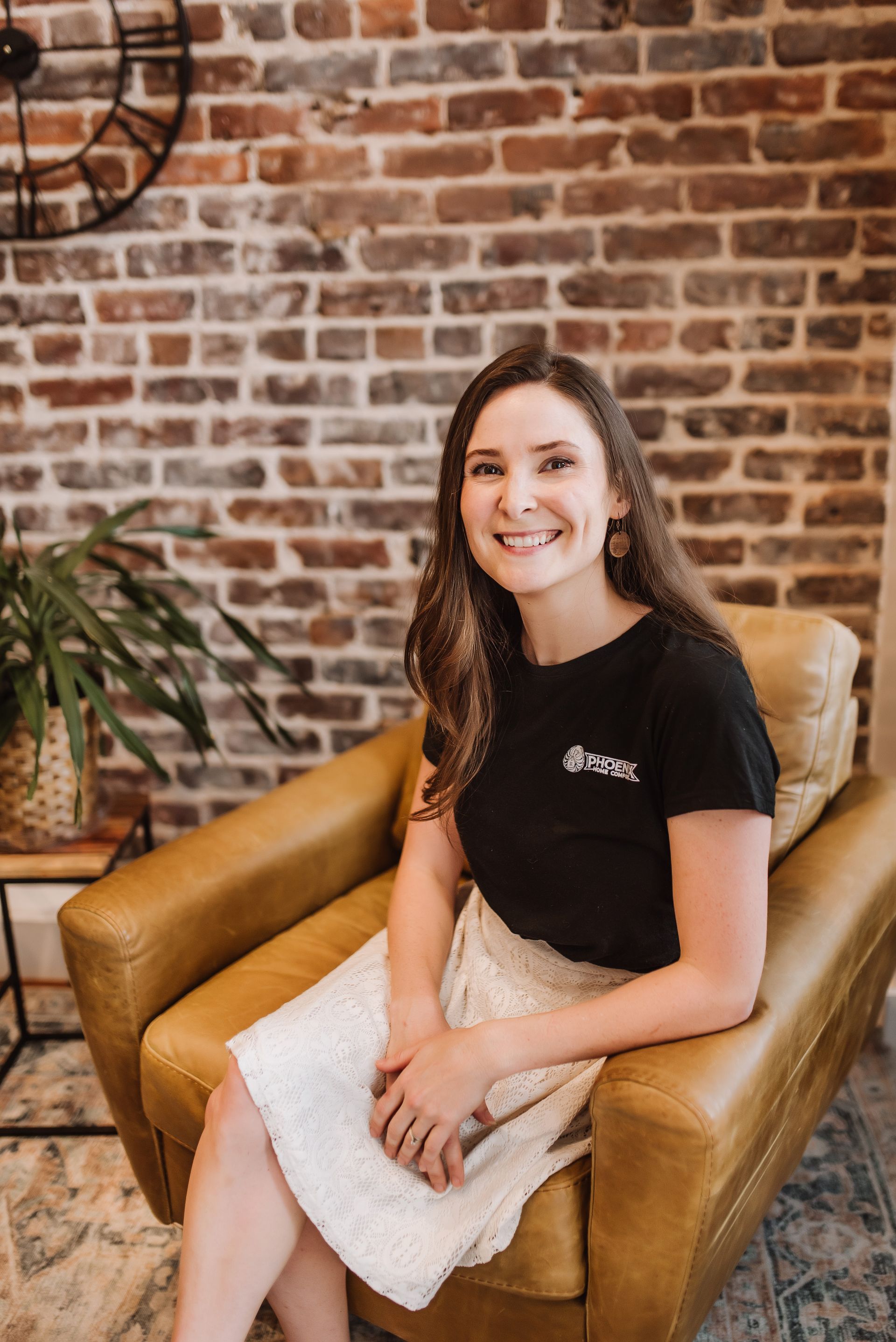 Woman smiling in a tan armchair against a brick wall, wearing a black shirt and white skirt.