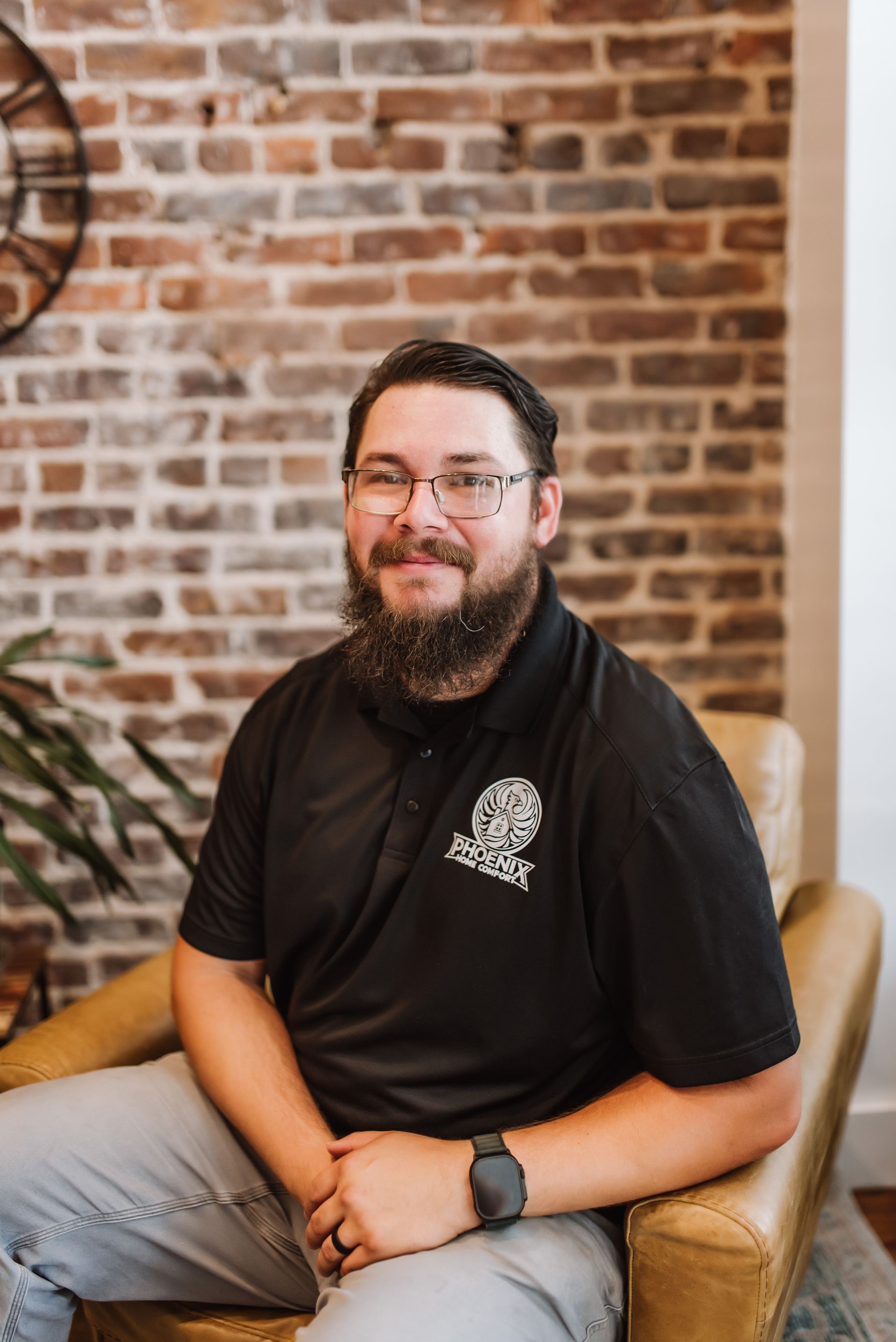 Smiling man in a black polo and jeans sitting in a chair against a brick wall.