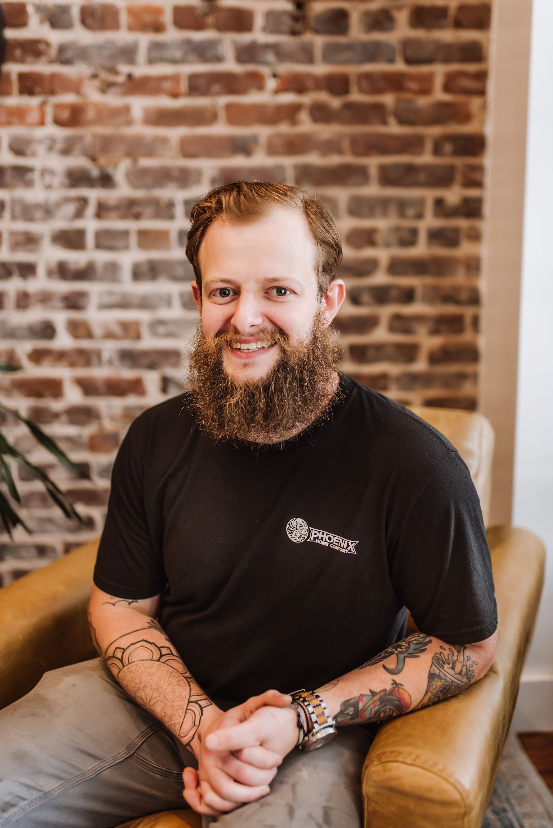 Smiling man with a beard in a black T-shirt, seated in a tan chair against a brick wall.