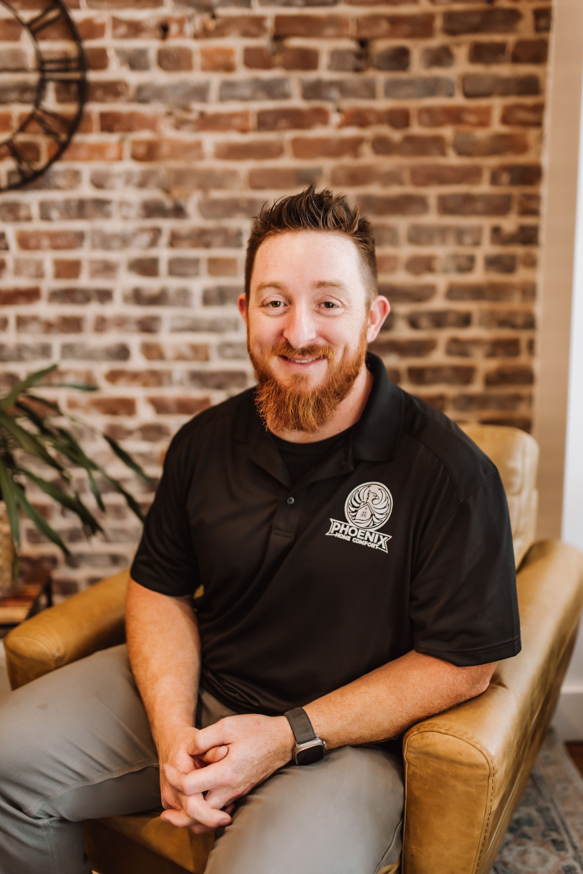 Man smiling in a black polo seated in a tan chair against a brick wall backdrop