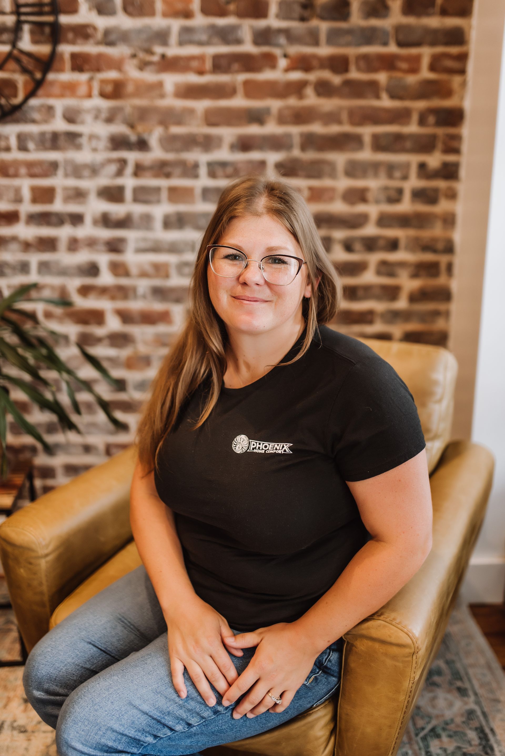 Woman sitting on a tan chair, smiling in front of a brick wall.