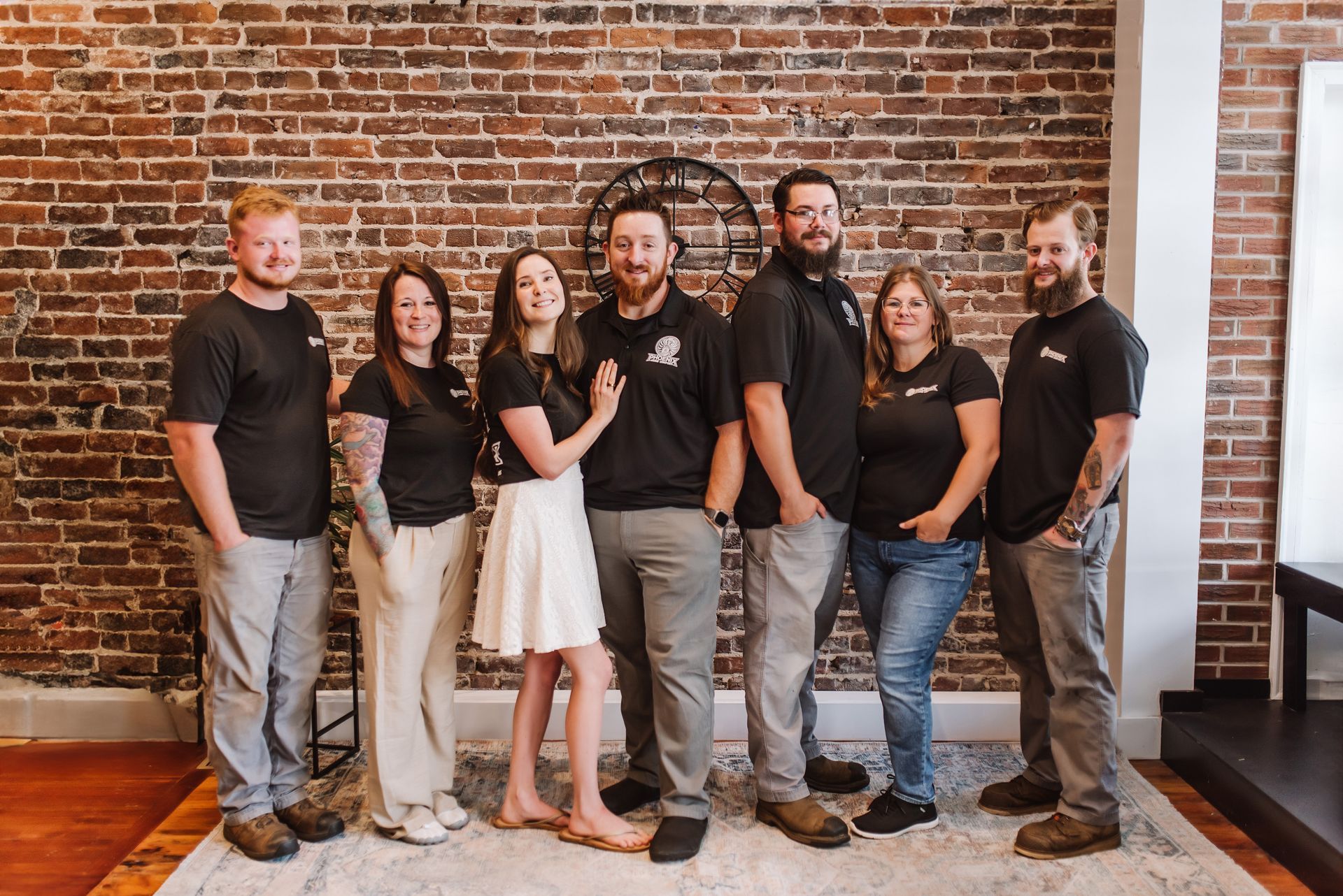 Group of seven people posing indoors against a brick wall, wearing matching black shirts and jeans.