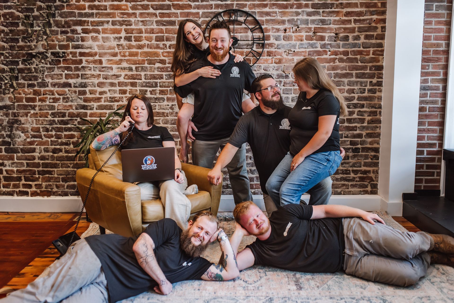 Group of six people lounging and smiling in a brick-walled room with a tan armchair and rug.