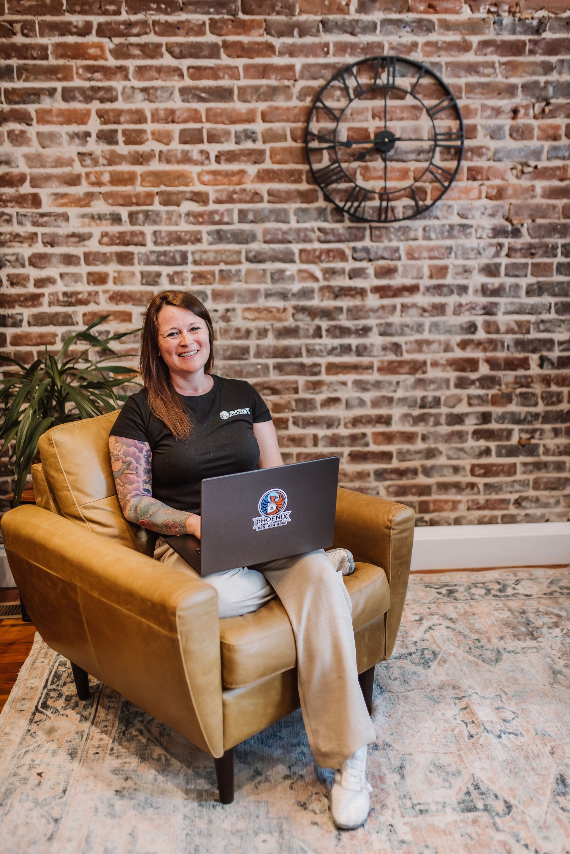 Woman sitting in a tan armchair with a laptop, in front of a brick wall and wall clock.