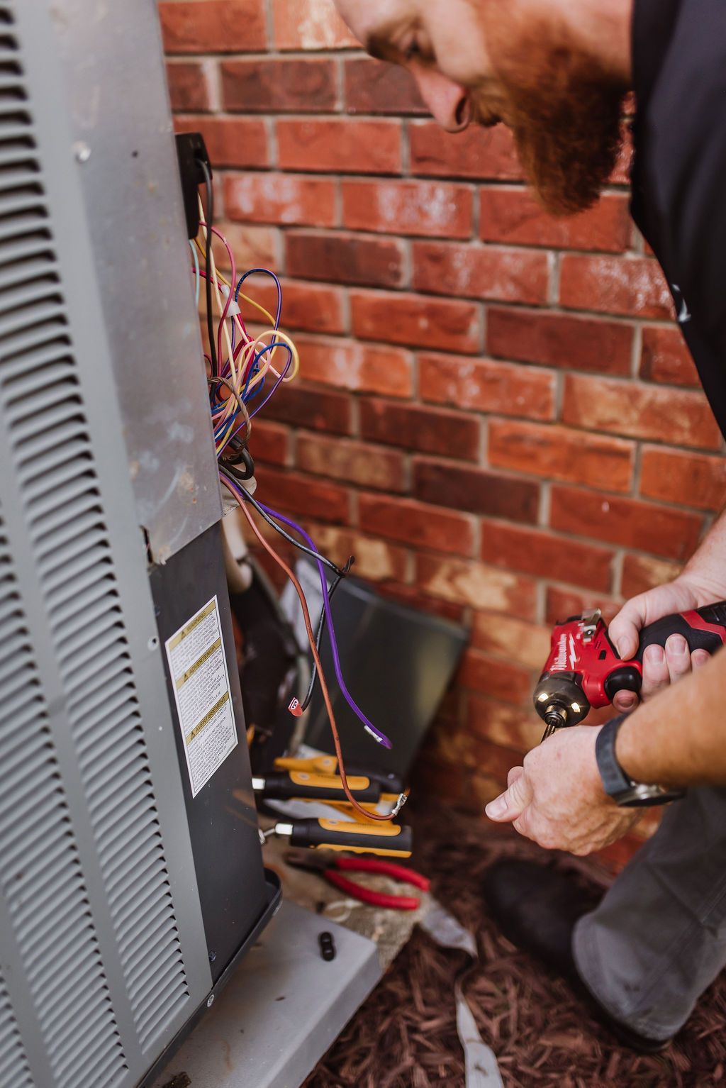 Technician working on exposed wires beside an outdoor HVAC unit against a brick wall