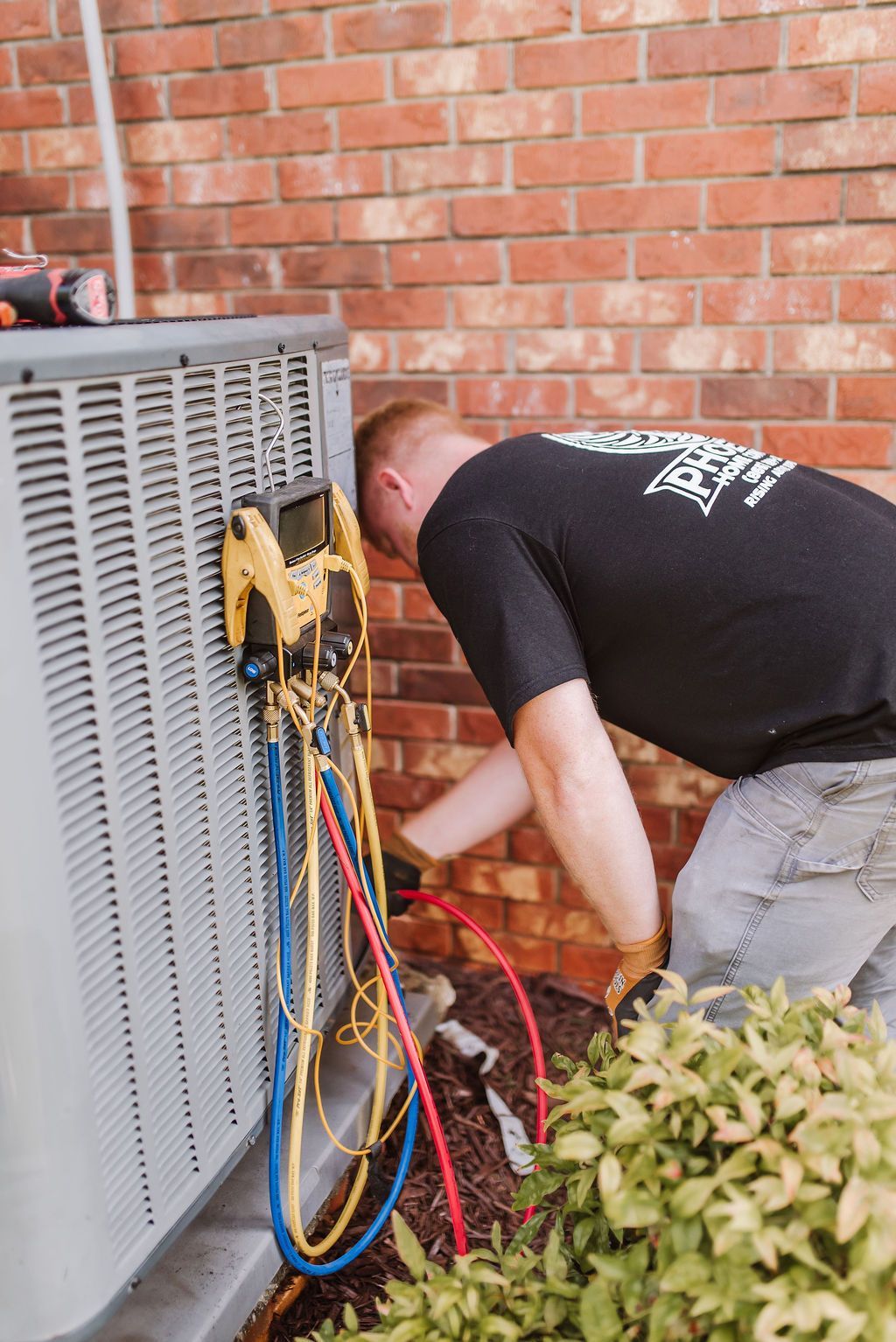 Technician kneels by outdoor HVAC unit, working on hoses and gauges against a brick wall.