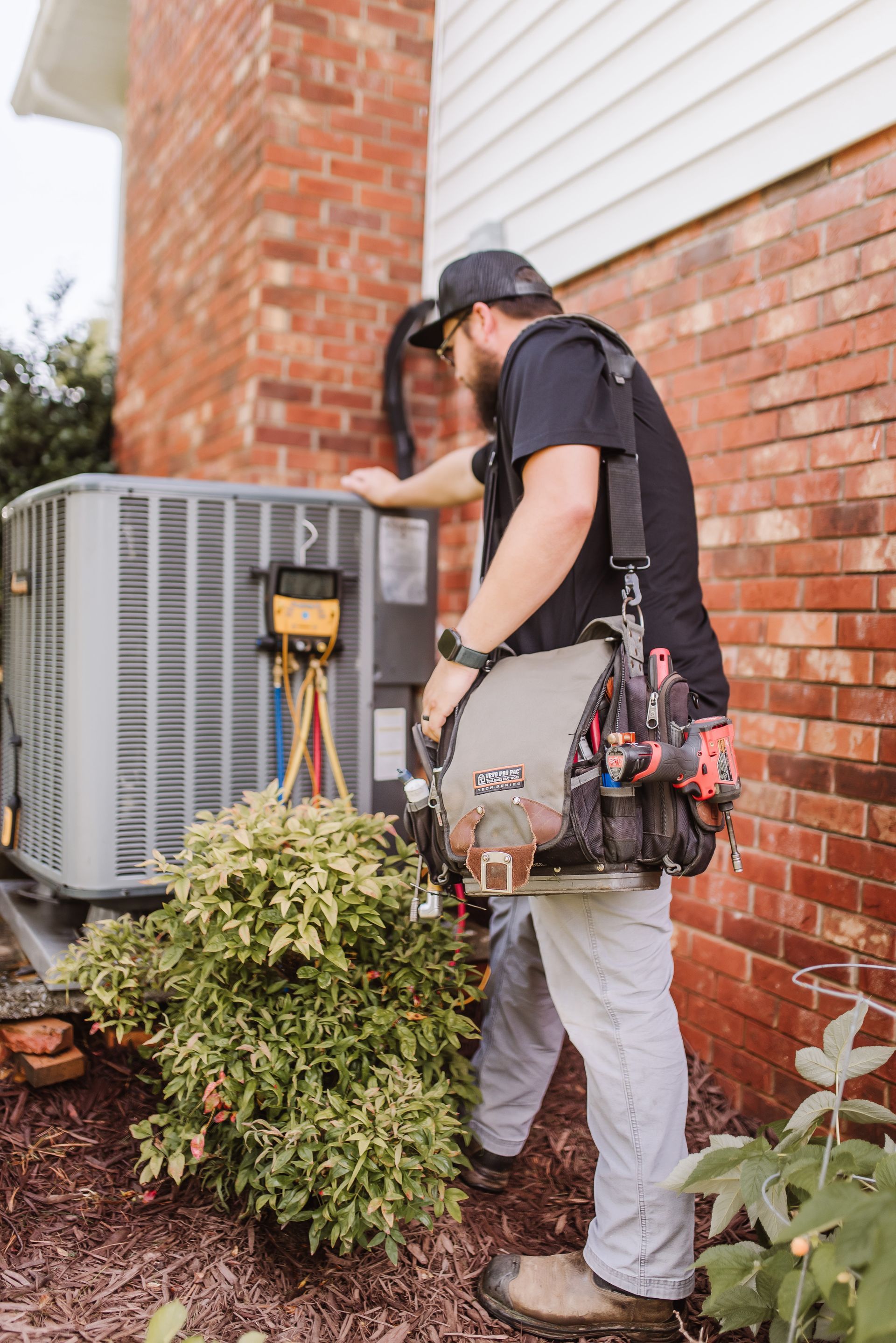 HVAC technician inspecting outdoor air conditioning unit beside a brick house