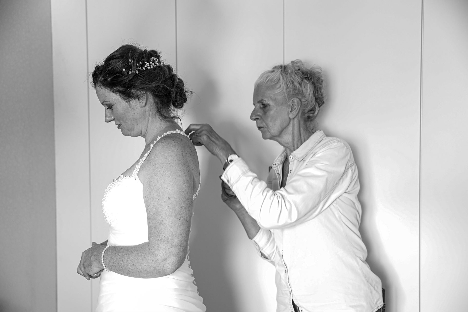 Een oudere vrouw maakt de achterkant van de witte trouwjurk van een jonge vrouw vast tegen een effen muur. Bruidsfotograaf.