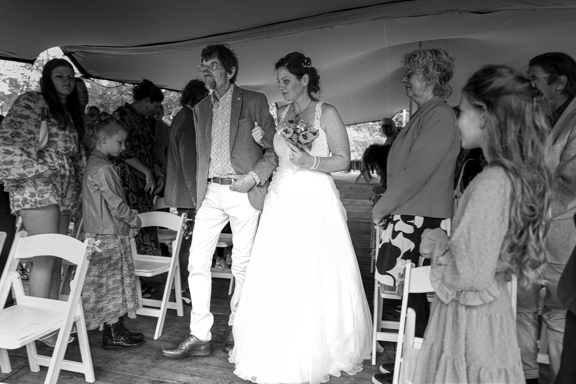 Bruid en bruidegom staan samen in een tent, omringd door gasten tijdens een huwelijksreceptie.