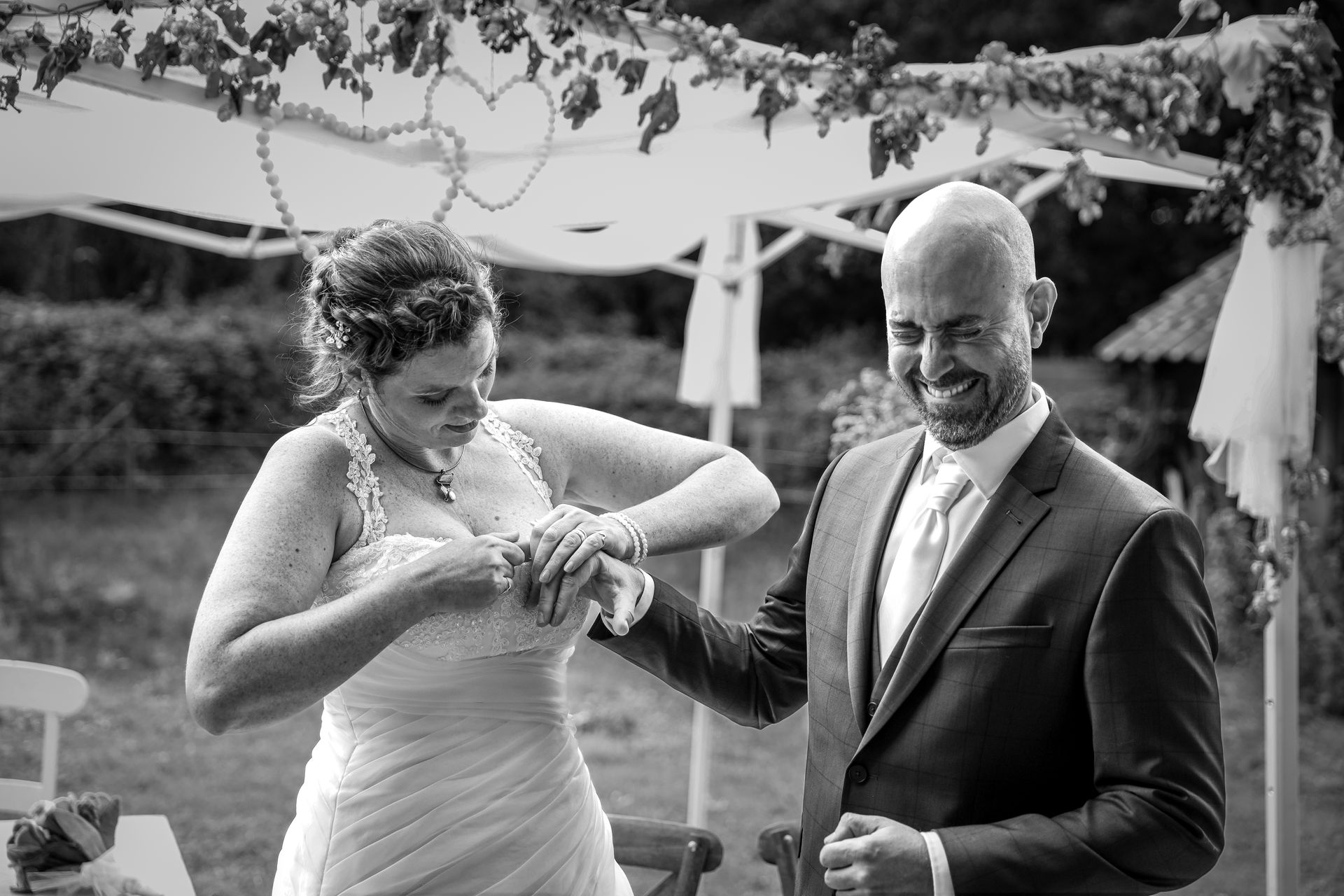 Bruid en bruidegom lachen terwijl zij zijn trouwring om schuift onder een versierde luifel buiten. Bruiloft fotografie.