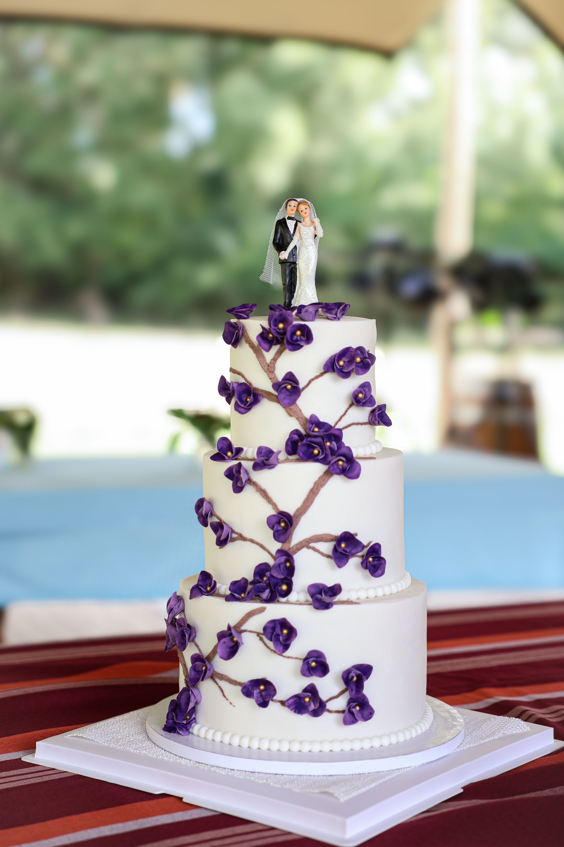 Een witte bruidstaart met drie lagen, versierd met paarse bloemen en een bruid en bruidegom als topper.