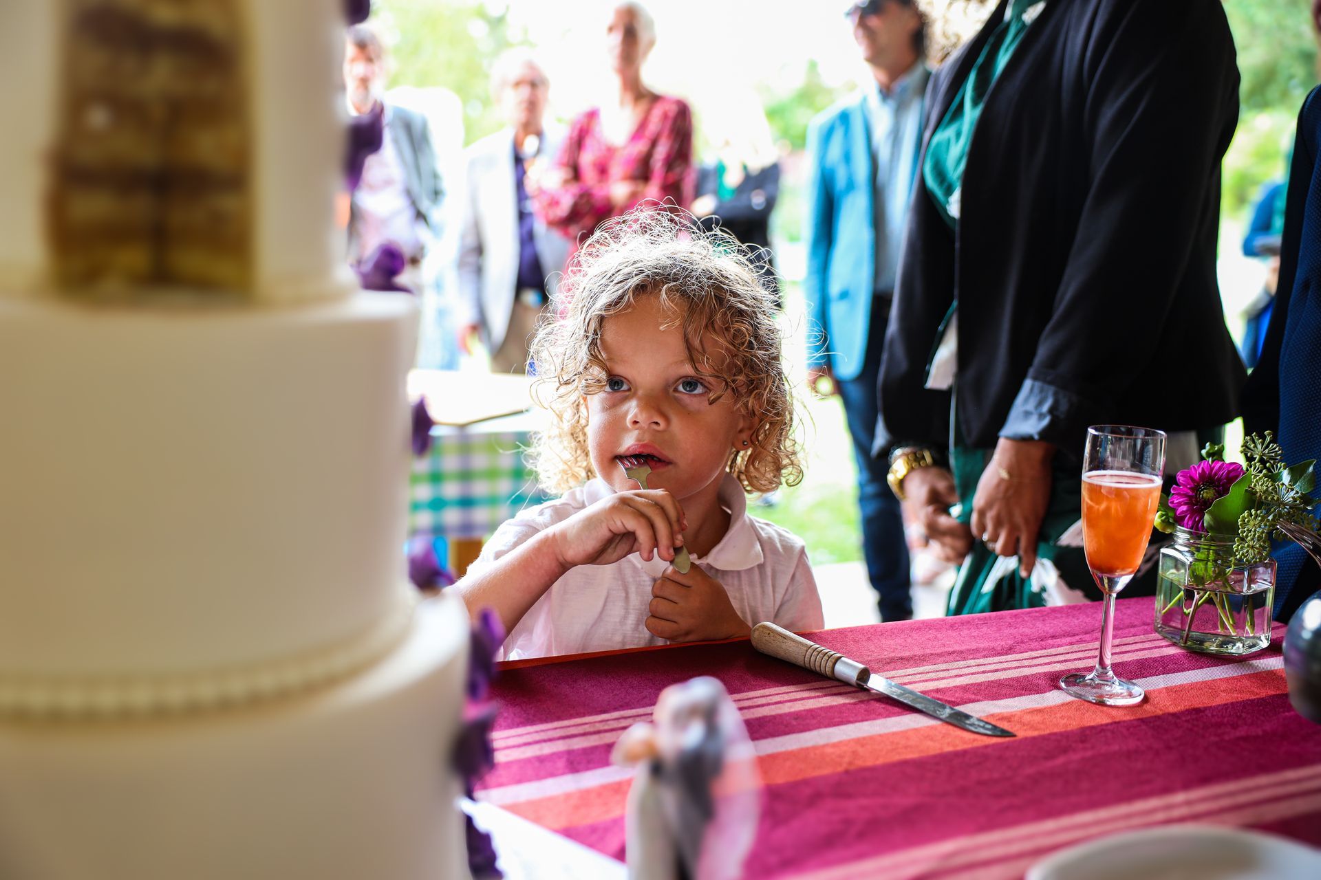 Kind zit aan een versierde tafel, kijkt voor zich uit met de handen bij het gezicht tijdens bruilift. Bruiloft fotograaf.