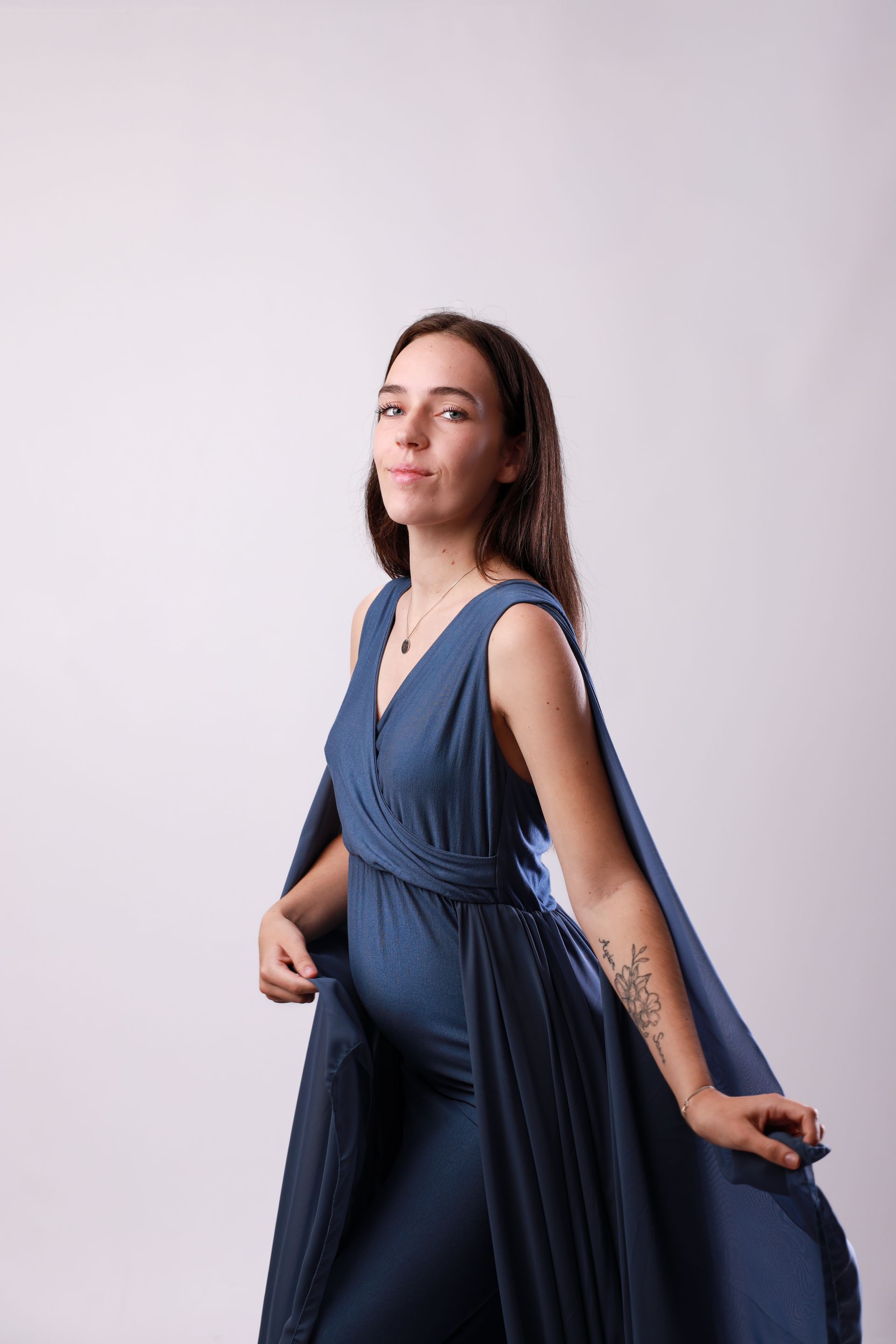Vrouw in een zwierige blauwe jurk poseert tegen een lichte, effen achtergrond. Zwangerschap fotograaf Hellevoetsluis.
