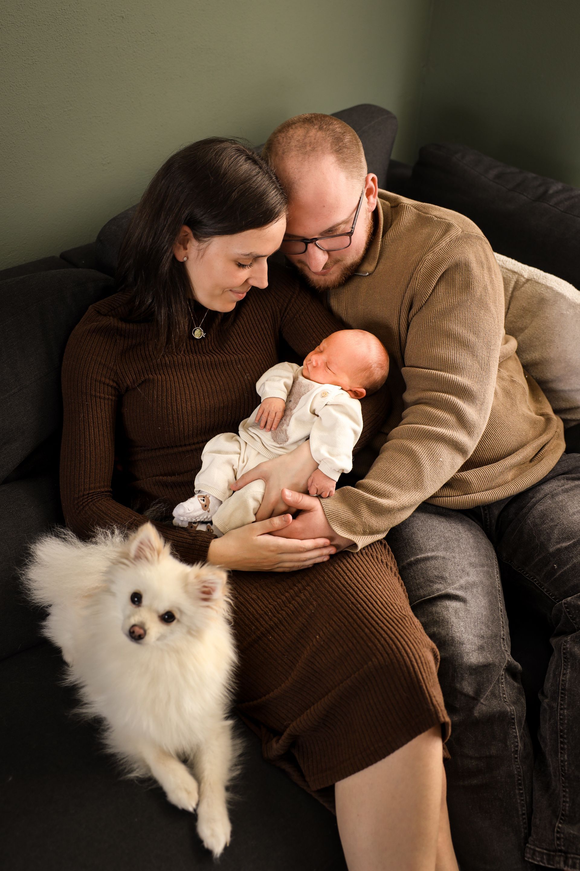 Ouders houden een slapende pasgeborene vast op de bank, met een pluizige witte hond ernaast. Newbornfotografie Hellevoetsluis