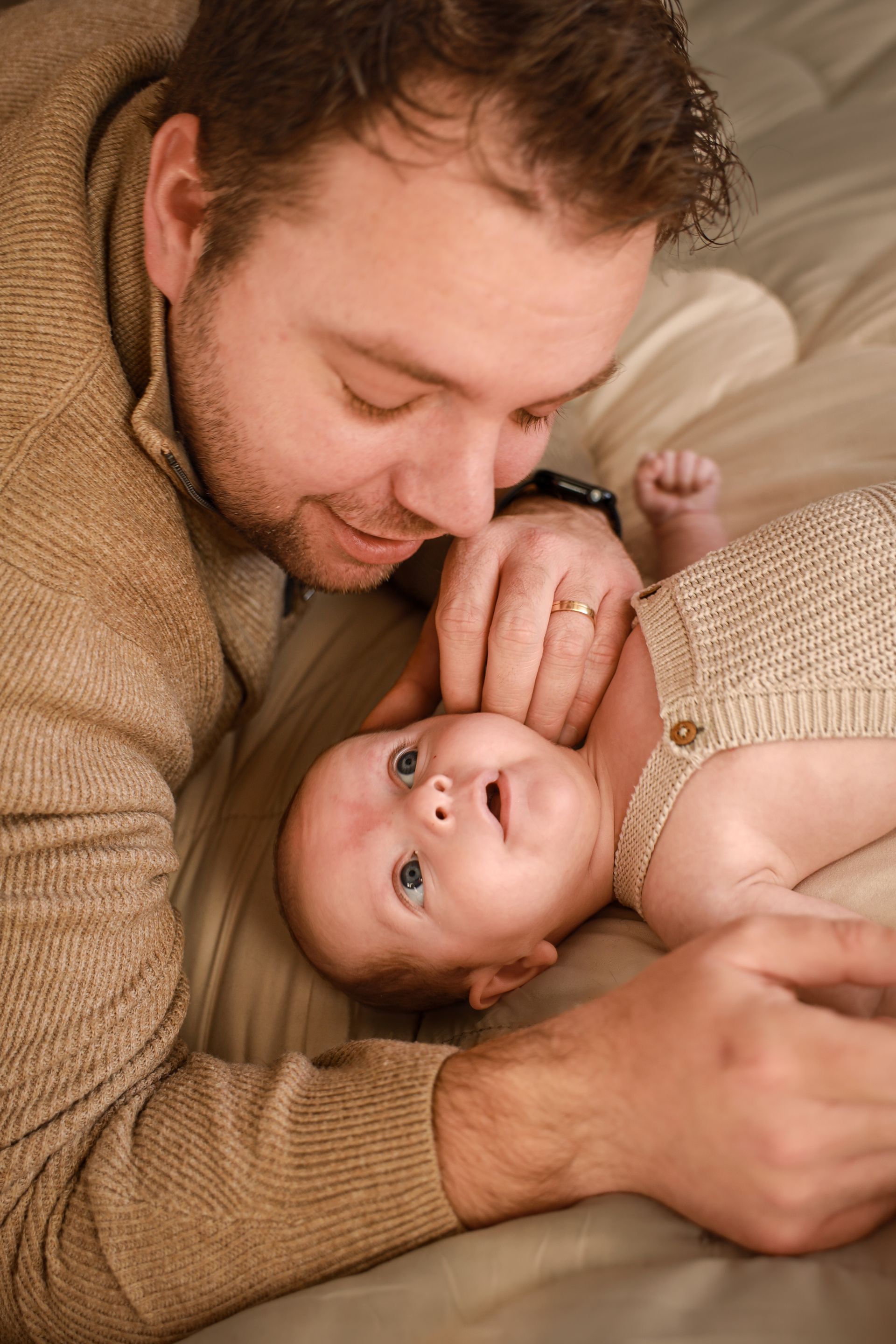 Een man ligt dicht bij een baby op een bed en raakt zachtjes het gezichtje van de baby aan. Newborn fotografie Spijkenisse.