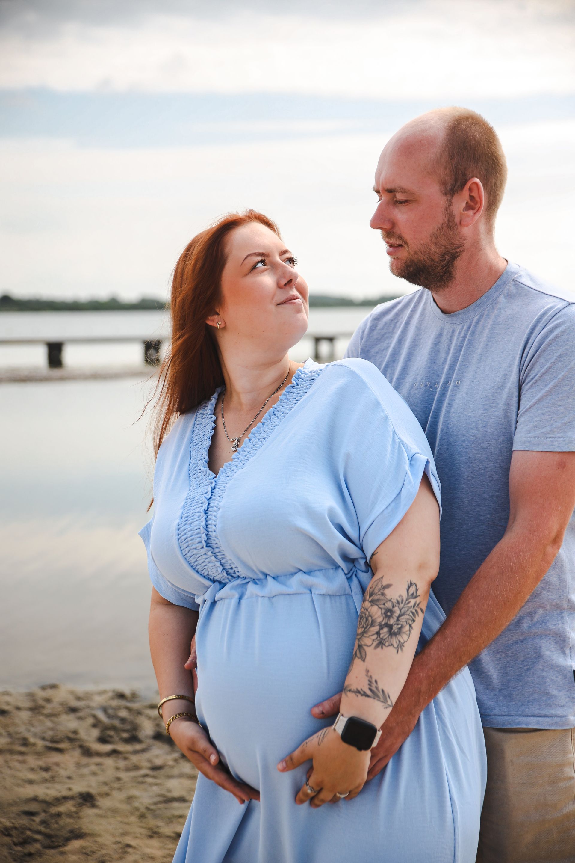 Een stel omhelst elkaar op het strand bij zonsondergang en lacht naar elkaar; babyshower fotograaf. babyshower fotoshoot.