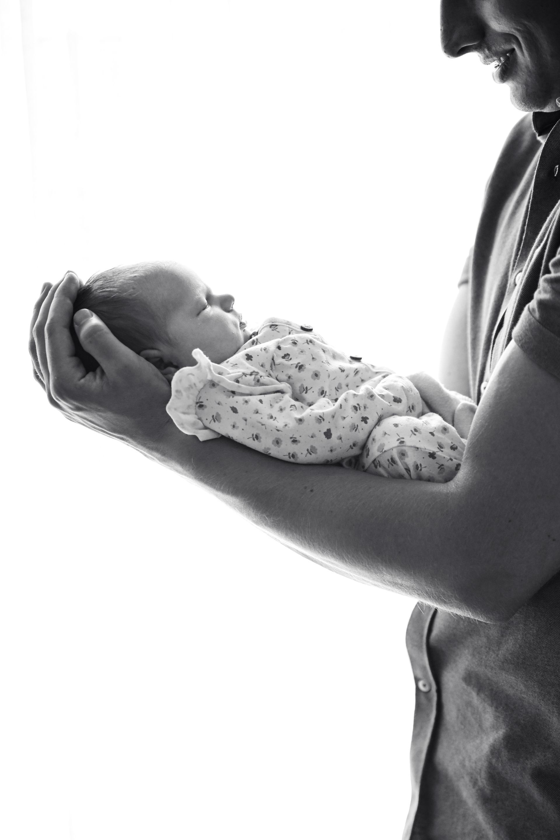 Een volwassene houdt een slapende baby in beide handen vast, gezicht aan gezicht, in zacht zwart-wit licht. Newborn fotograaf