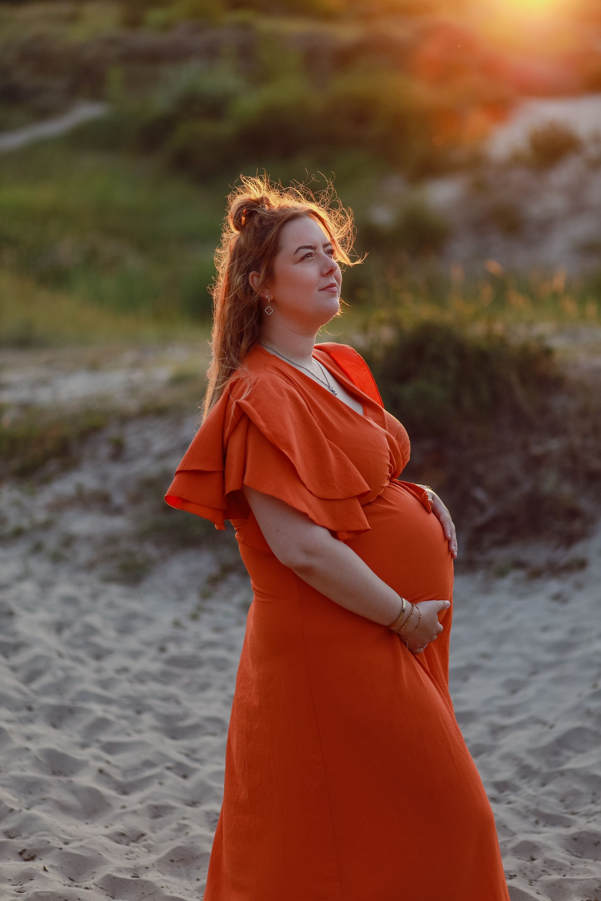 Vrouw in een feloranje jurk staat op een zandstrand bij zonsondergang. Zwangerschapsfotografie Voorne Putten, Zuidland.