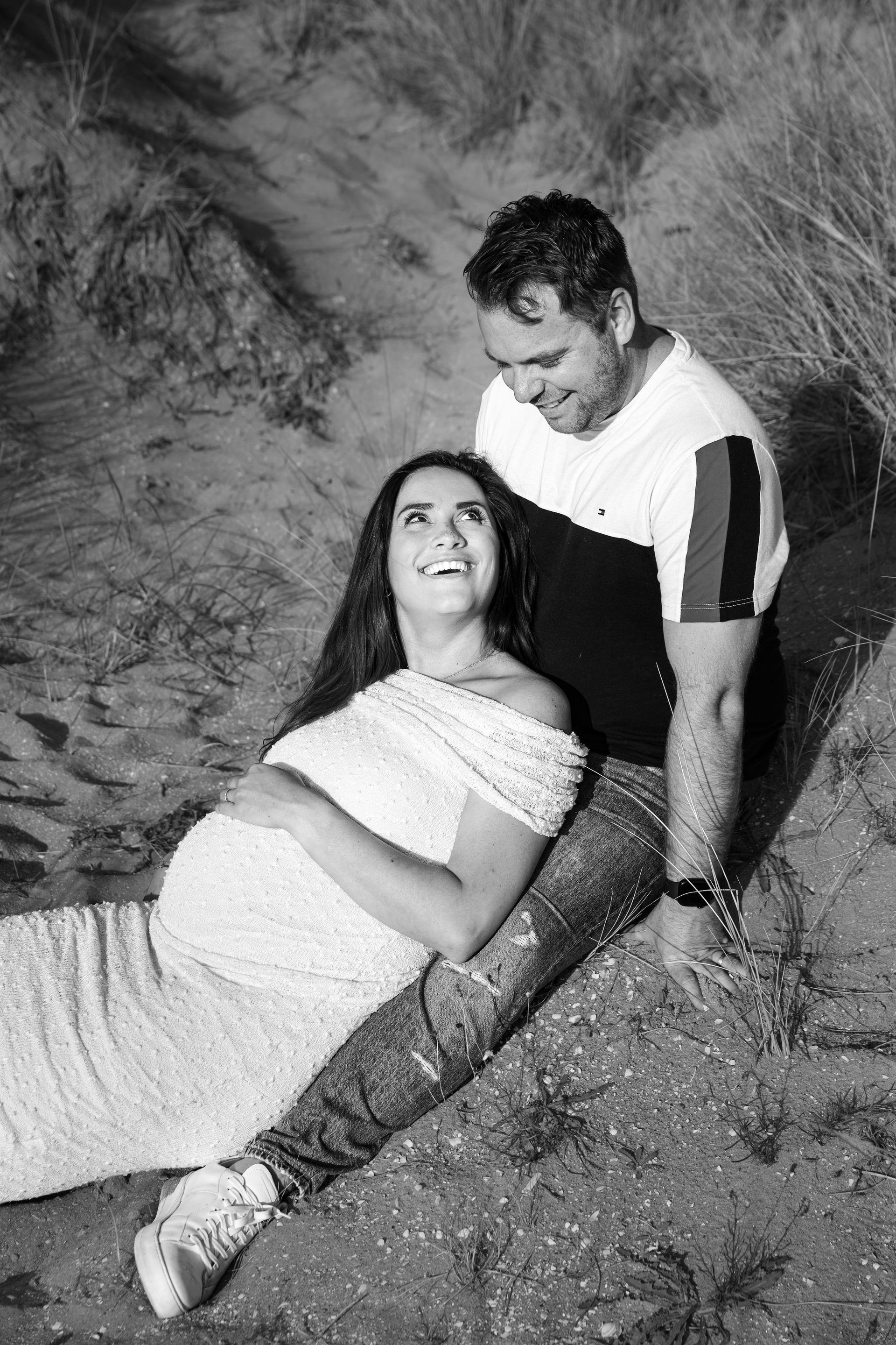 Een stel zit op een zandhelling, de vrouw leunt achterover tegen de man in een gestreept shirt. Zwangerschapsfotograaf.