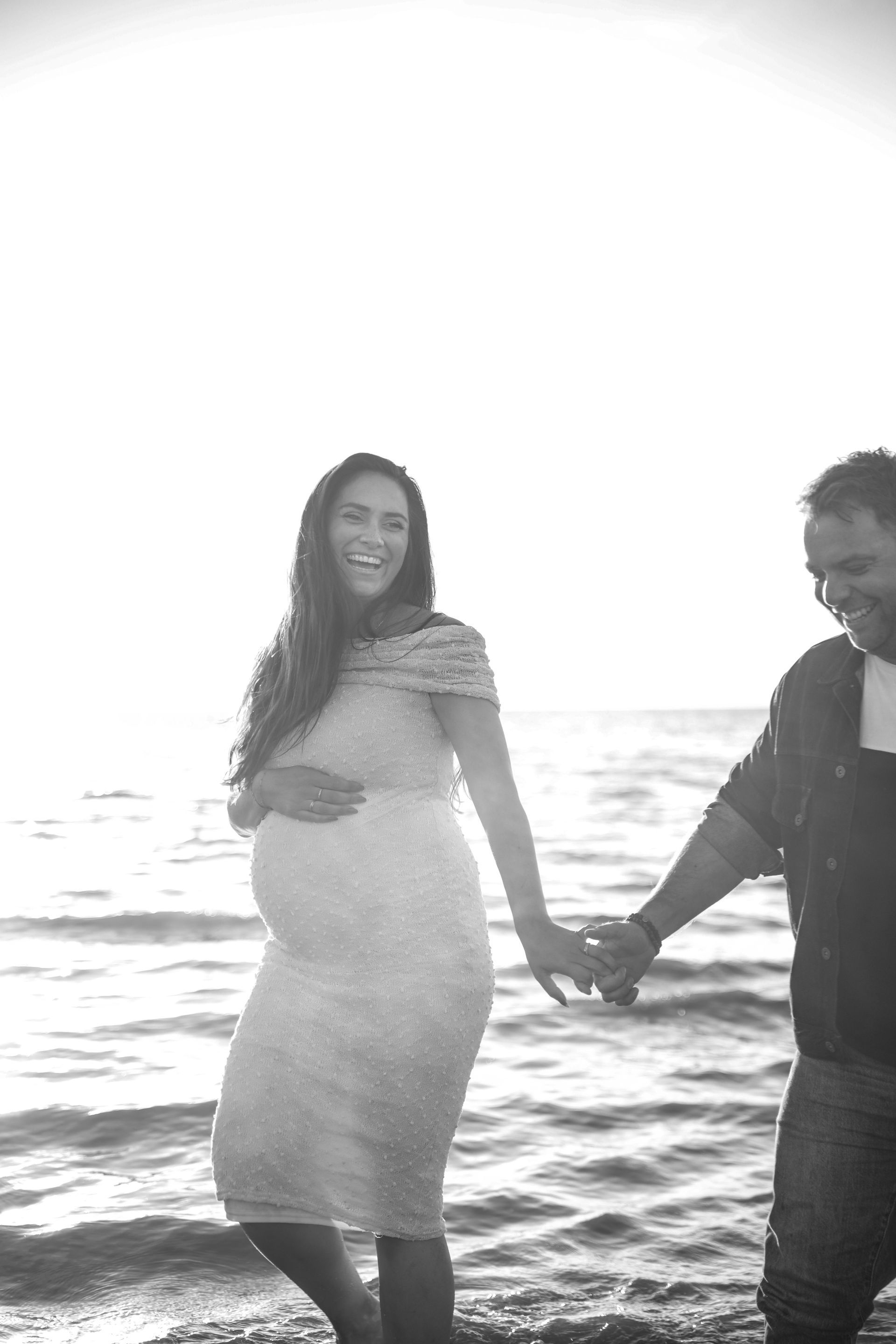 Een zwangere vrouw houdt de hand van een metgezel vast terwijl ze in het ondiepe zeewater op het strand staat.