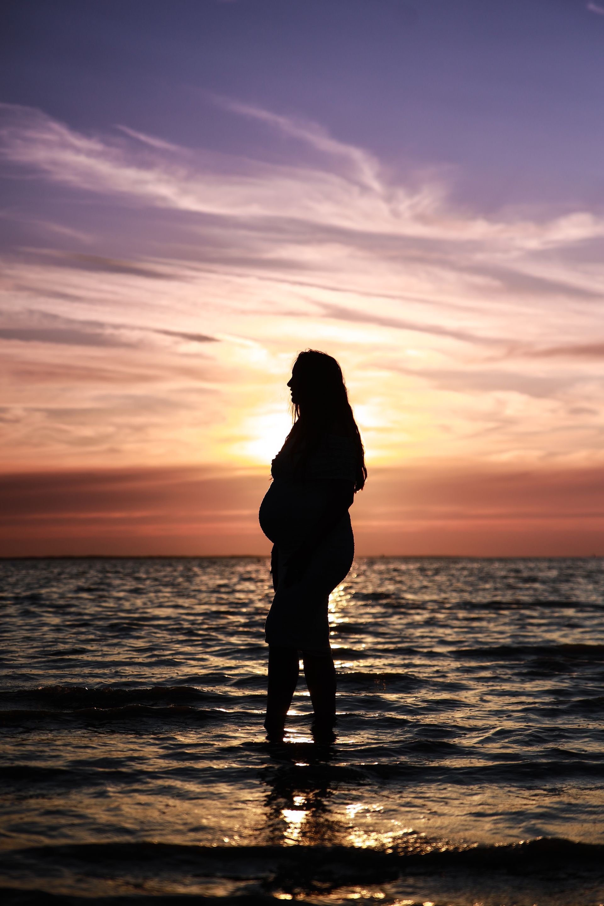 Silhouet van een persoon die in ondiep oceaanwater staat bij zonsondergang, met een oranje en paarse lucht. Zwanger fotoshoot