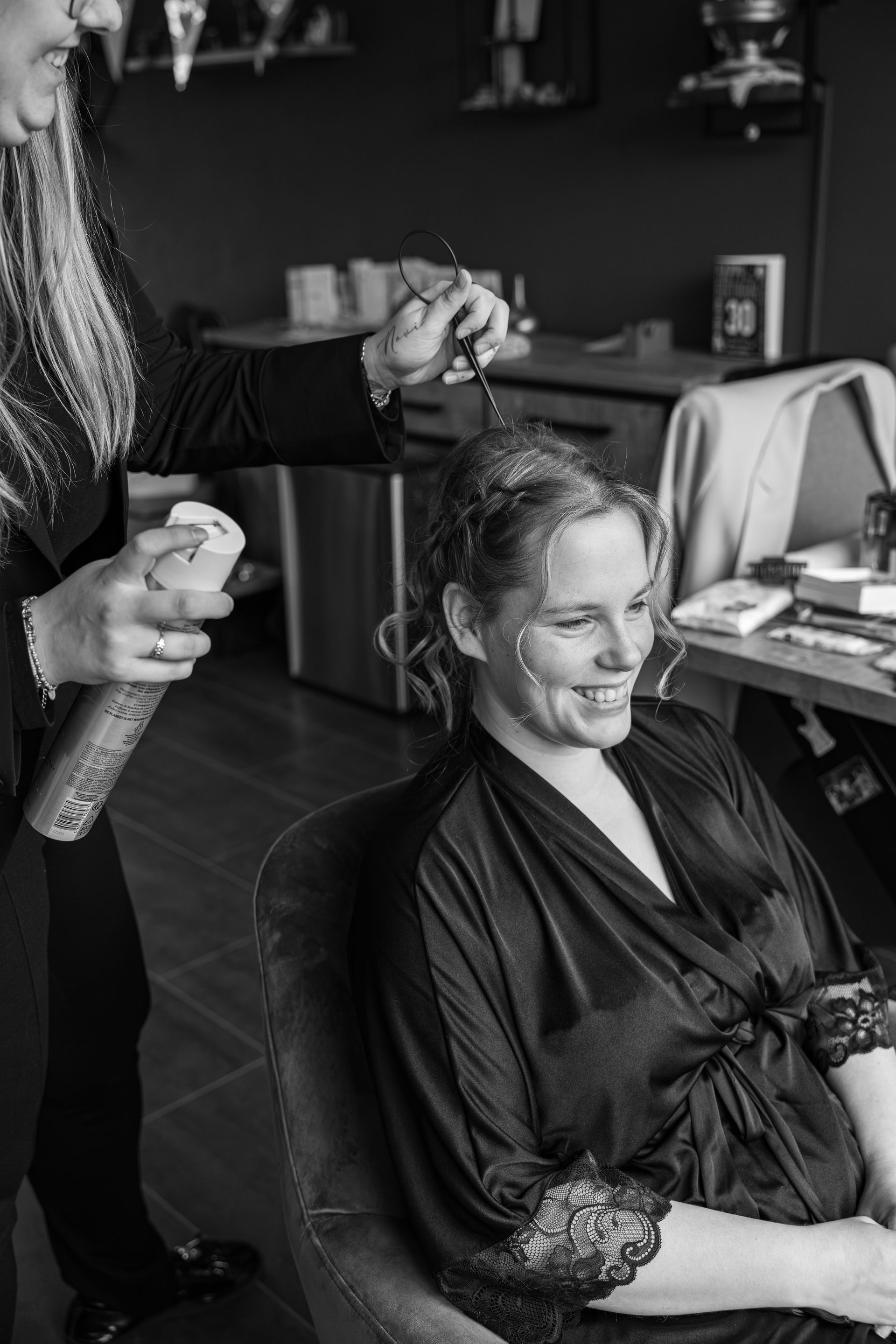 Een persoon krijgt haarspray door een stylist terwijl ze in een kappersstoel zit en lacht. Goedkope bruiloft fotograaf.